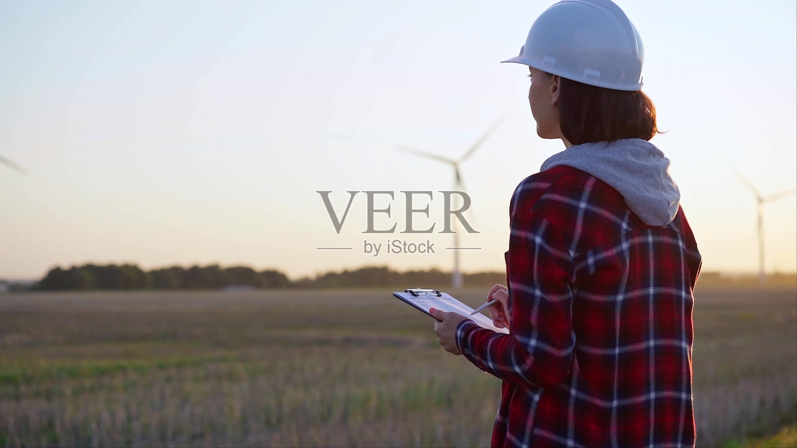 身穿白色安全帽的成年女性工程师在风力发电机旁的现场用夹板做笔记，背影在日落时分。照片摄影图片