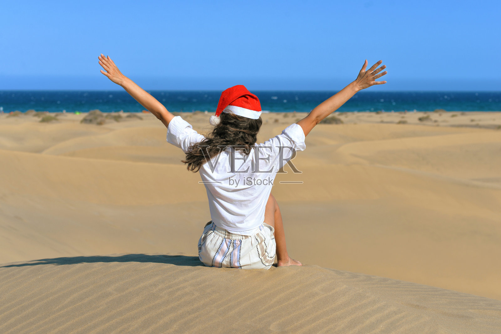 一个女孩在沙丘上戴着圣诞帽，双手举起。背影。热带国家海边的圣诞假期概念。位于大加那利岛马斯帕洛马斯的海边沙丘。照片摄影图片