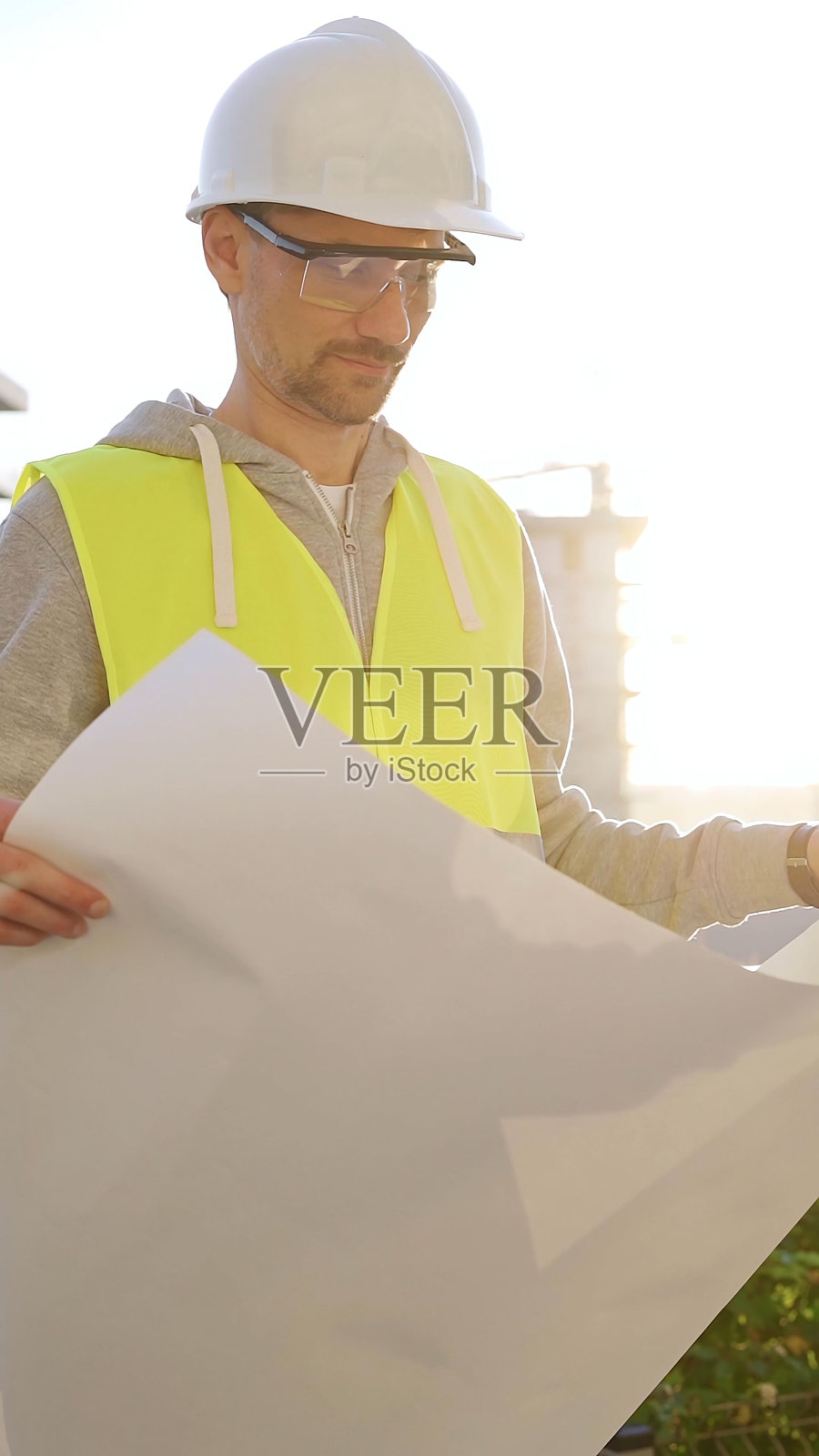 一名建筑师或工程师穿着安全帽和马甲，手持蓝图，面朝日出时的建筑工地，正面垂直视角。建筑概念照片摄影图片