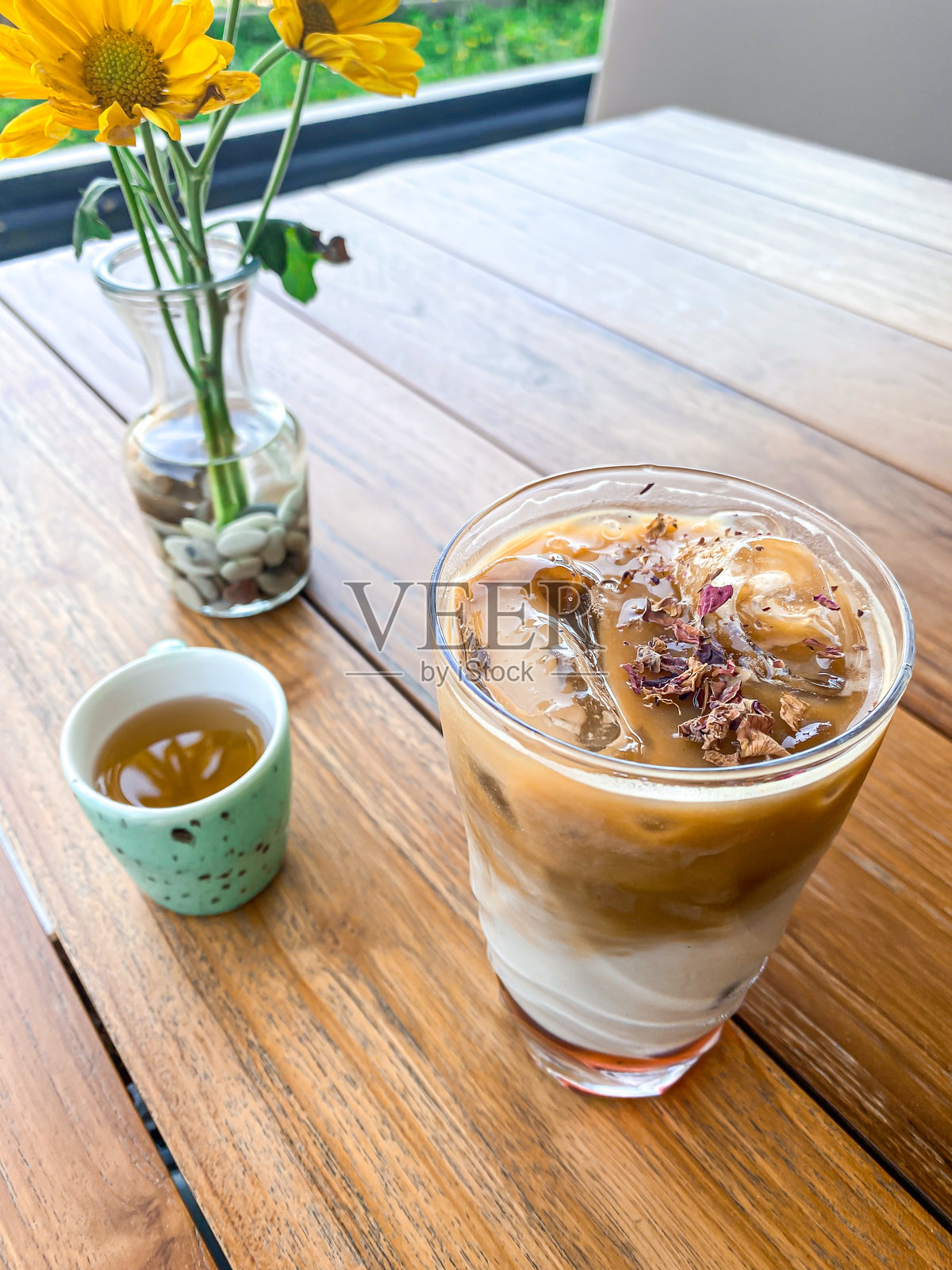 透明玻璃杯中的冰卡布奇诺咖啡，上面撒有干玫瑰花瓣，旁边是液体糖杯和花瓶，放在木桌上。咖啡拿铁或咖啡牛奶饮品在杯子里。照片摄影图片