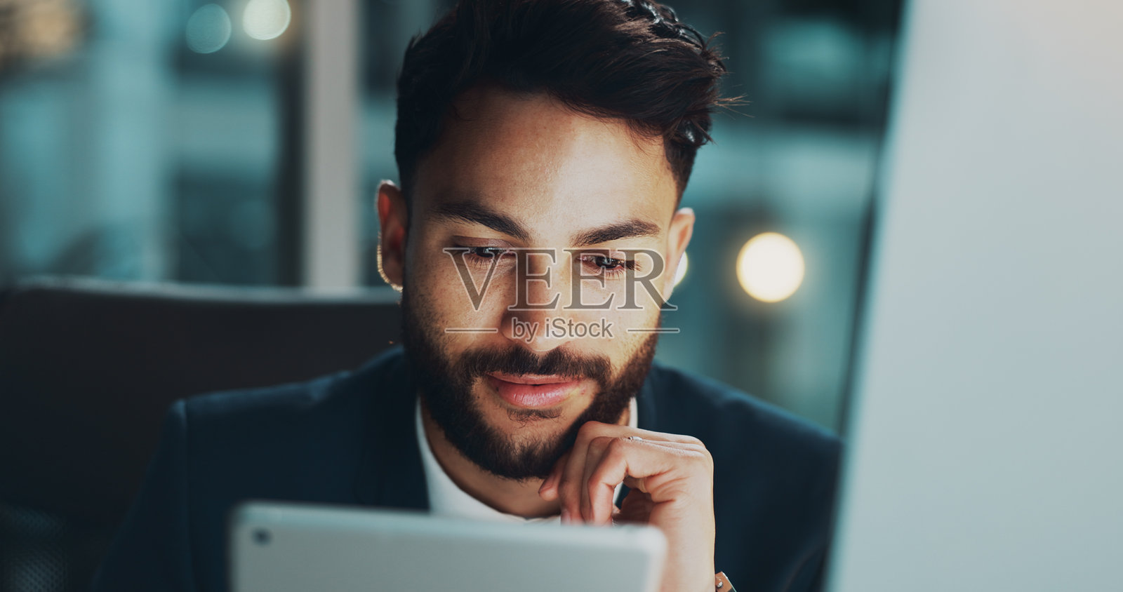 夜晚，办公室里的男人微笑着阅读平板电脑，正在为网络项目创意制定商业计划。他在科技公司加班，进行研究，使用数字应用程序处理电子邮件、报告或在线日程。照片摄影图片