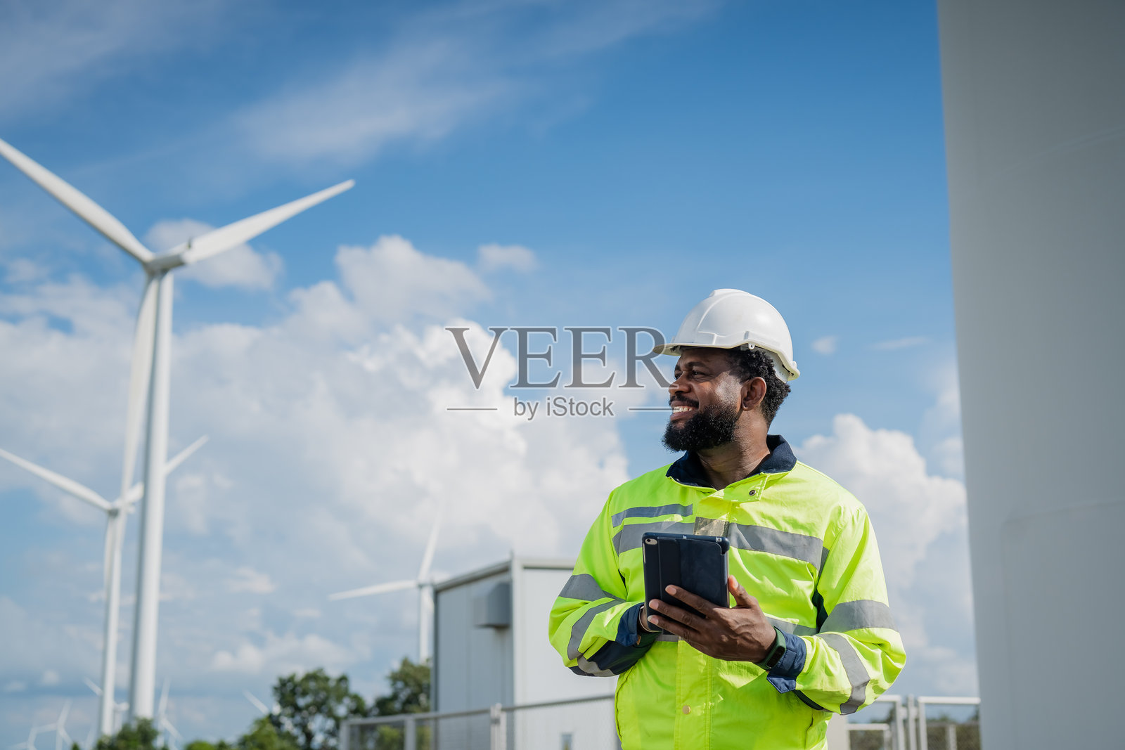 一名穿着黄色安全夹克的男子站在风力涡轮机旁，手持平板电脑。照片摄影图片