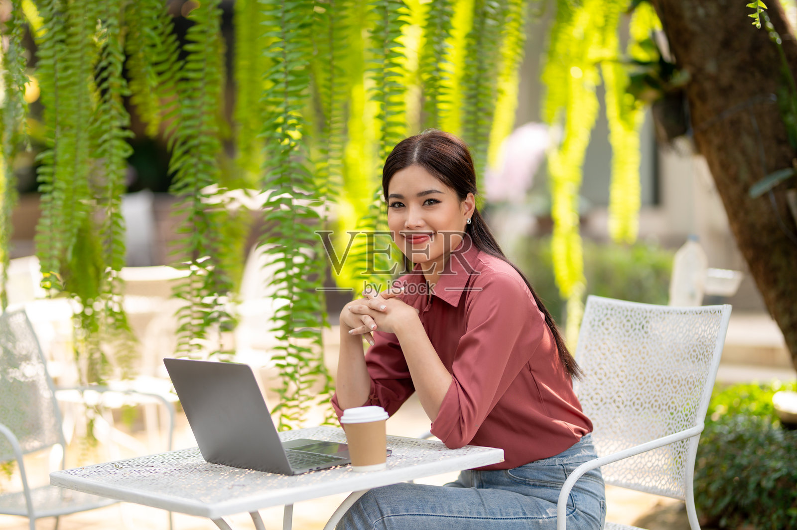 年轻的职业女性坐在舒适的藤椅上使用笔记本电脑，周围环绕着郁郁葱葱的绿植。照片摄影图片