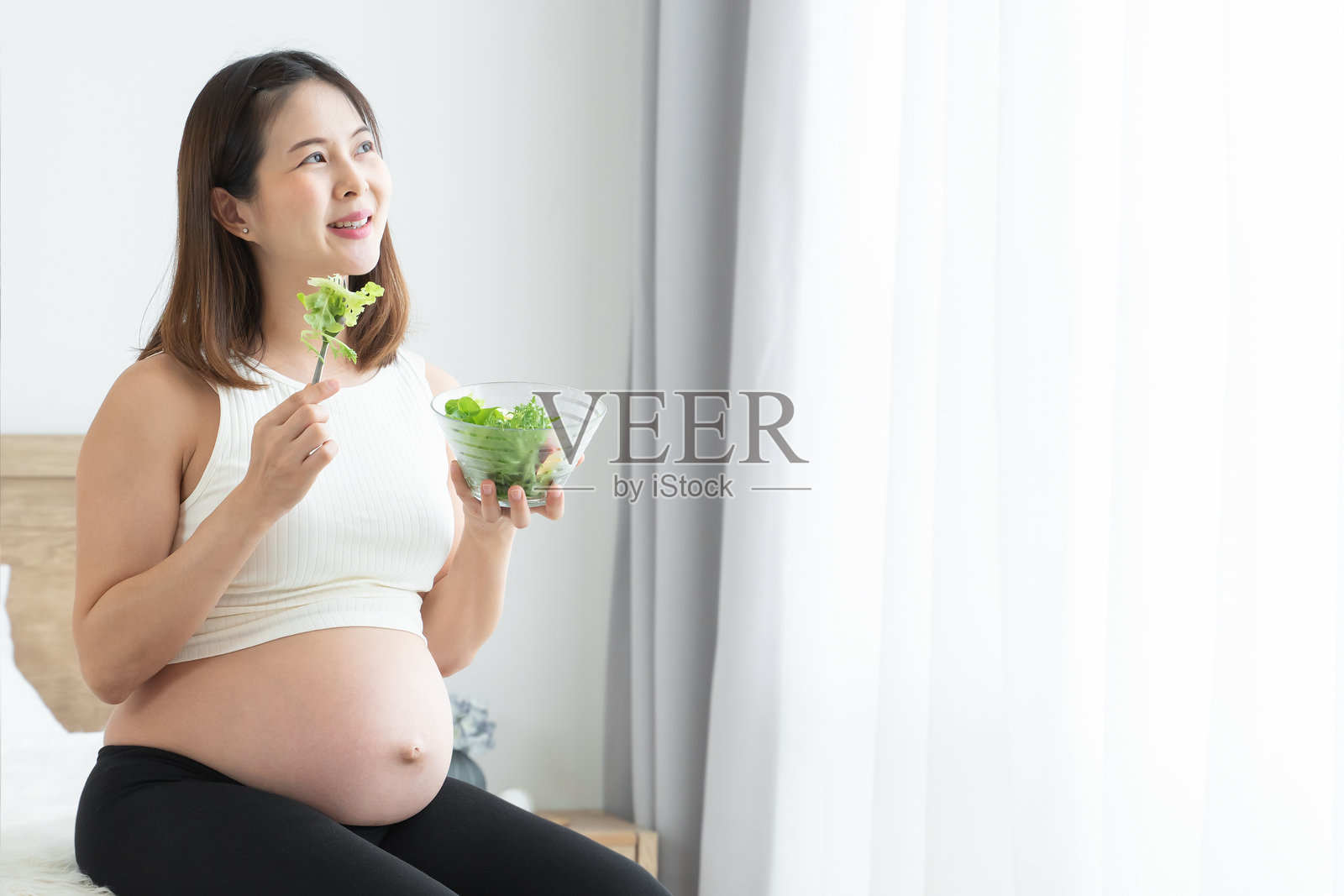 年轻的孕妇坐着，手里拿着一碗沙拉和叉子。她微笑着，望向卧室窗外。在家中，这位美丽的亚洲母亲正在享用新鲜的蔬菜沙拉，以保持健康。照片摄影图片
