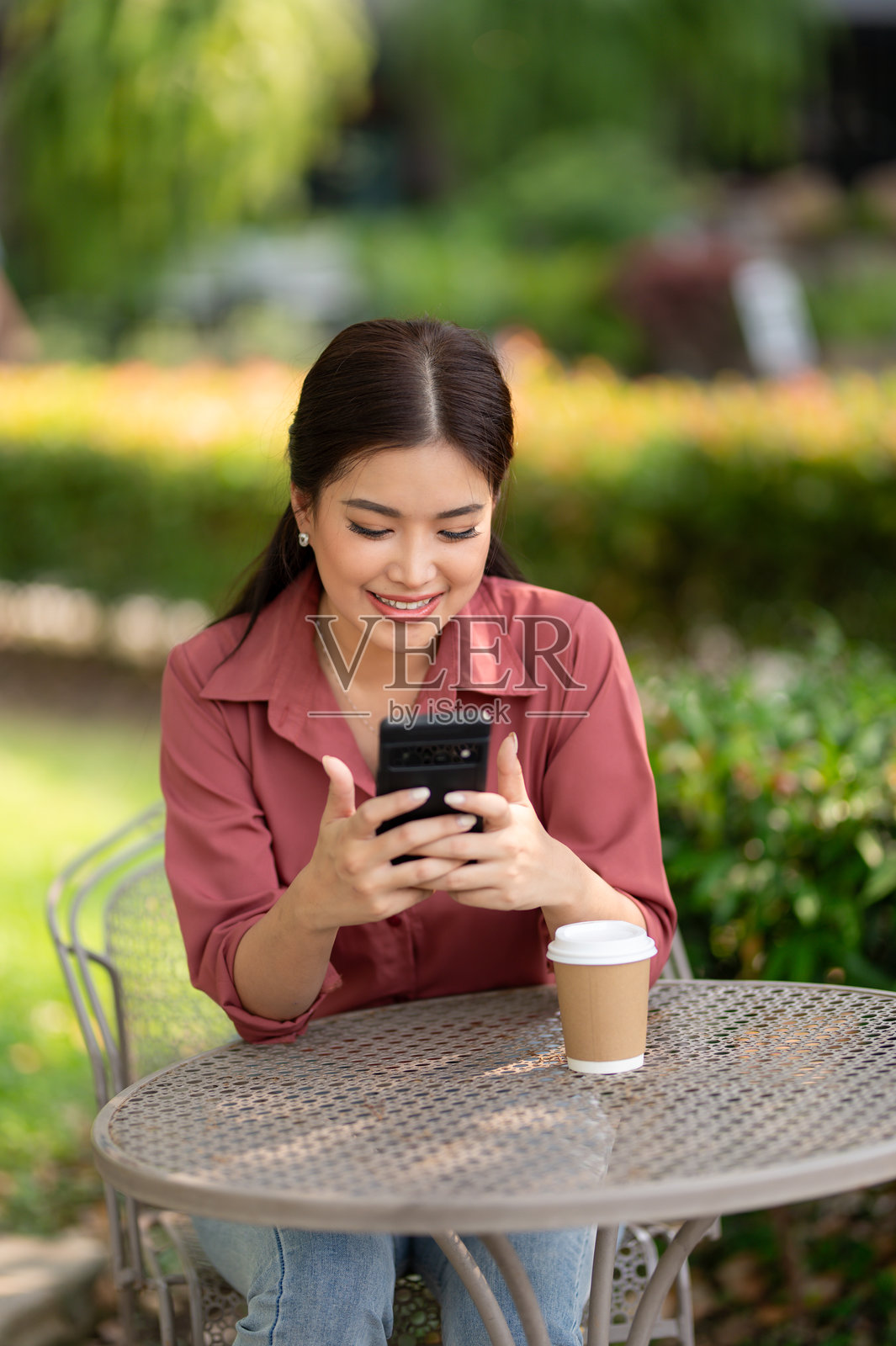 一位开心的年轻女性在宁静的花园咖啡馆享受咖啡时查看她的智能手机。照片摄影图片