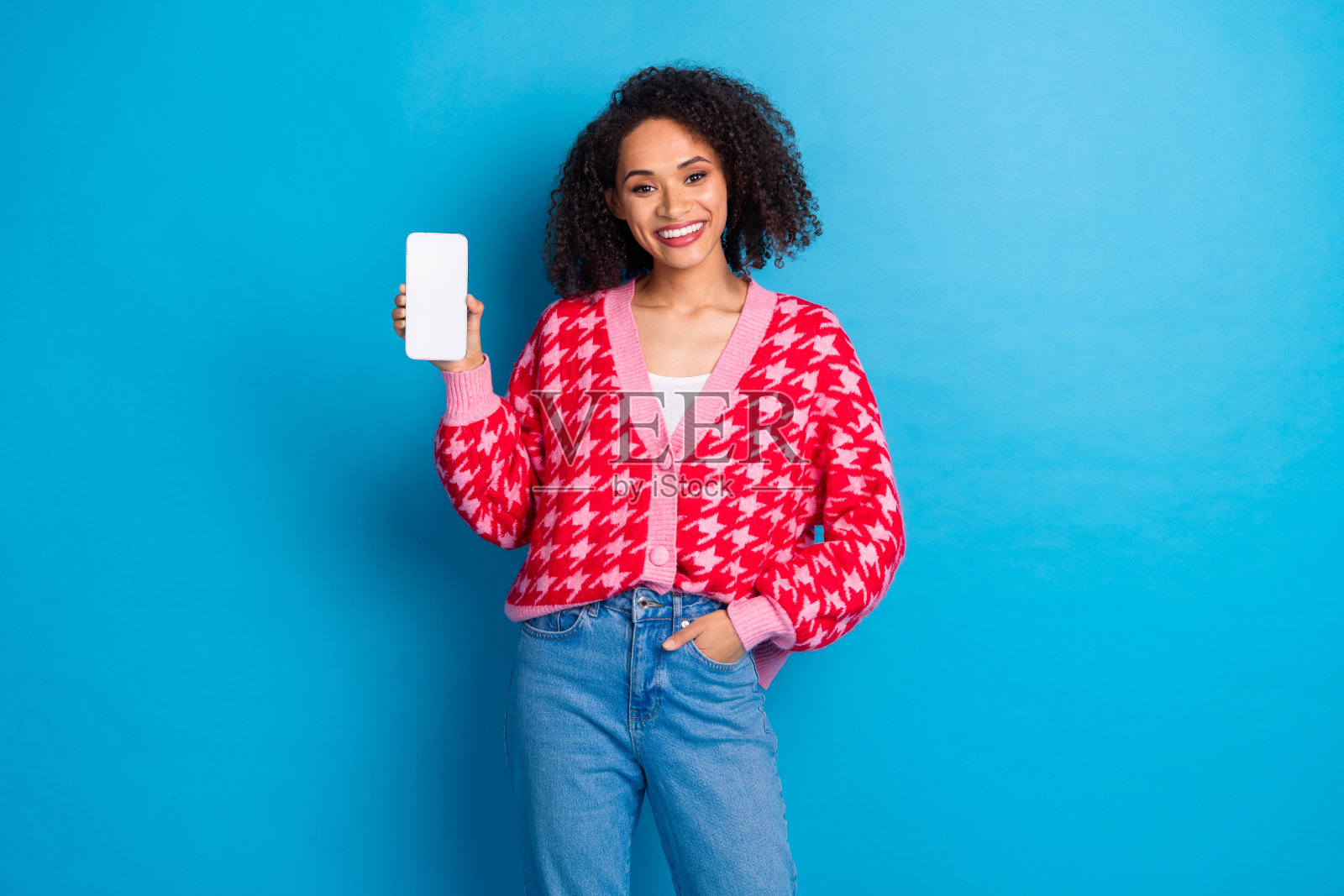 微笑的迷人女孩，卷发，穿着红色时尚开衫，手持手机，背景为蓝色。照片摄影图片