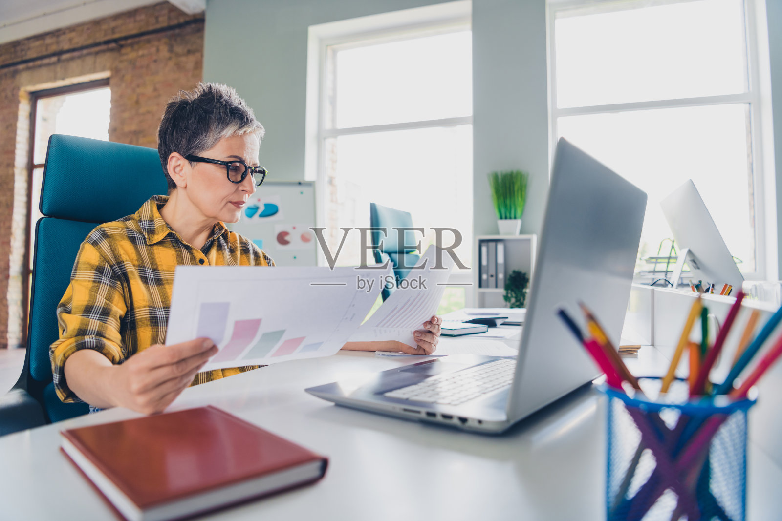 穿着格子黄色衬衫的漂亮老年女性在明亮的办公室中工作图表，展现出成功的合作氛围。照片摄影图片
