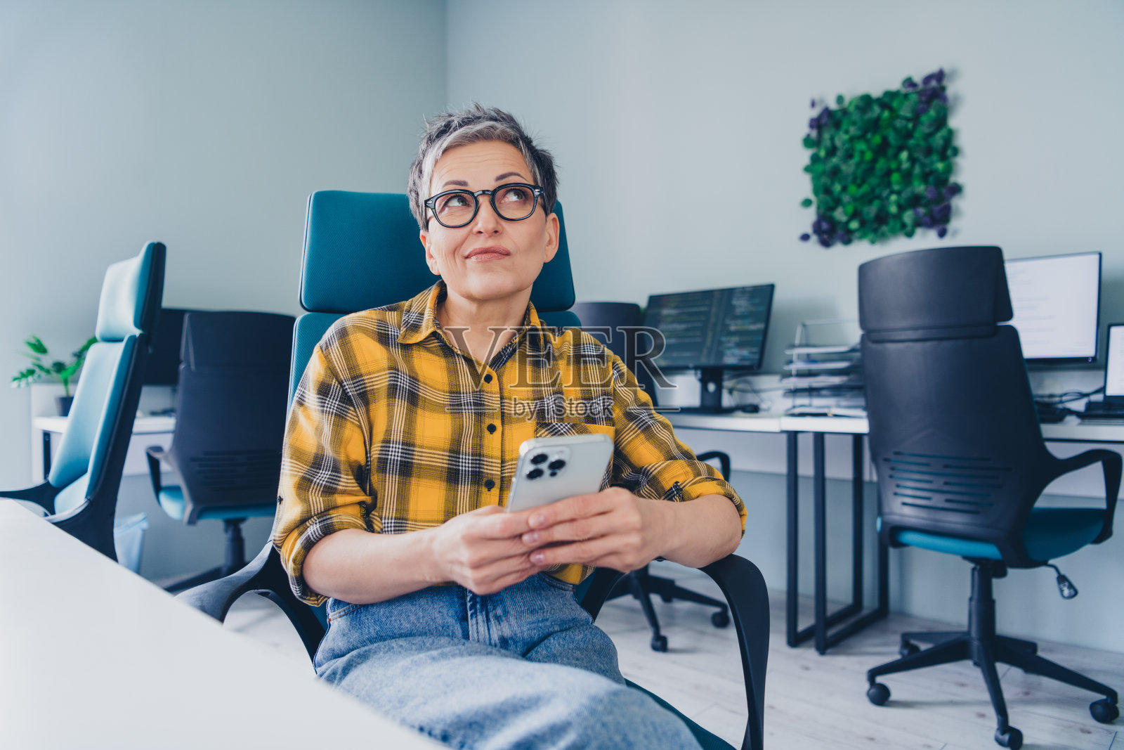 可爱的退休女士怀抱设备，怀疑地看着空旷的空间，穿着舒适的黄色格子衬衫，坐在初创公司的办公室内。照片摄影图片