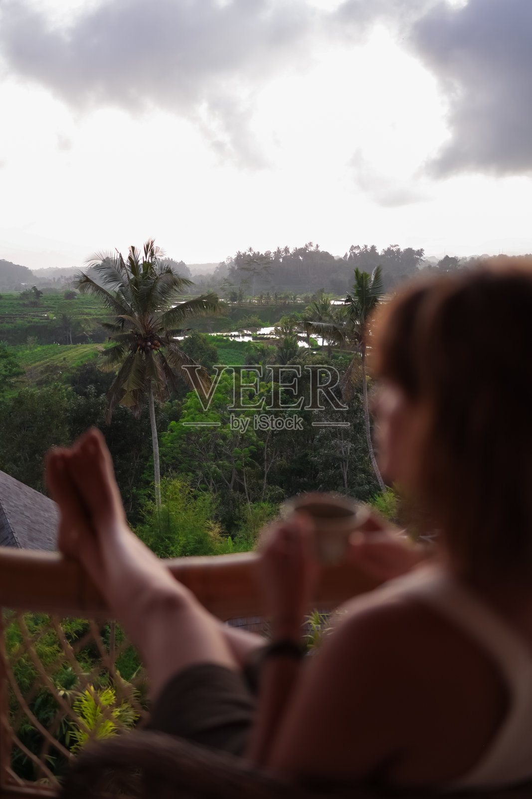 女人在印尼巴厘岛的竹屋阳台上欣赏宁静的早晨美景，俯瞰郁郁葱葱的田野，悠闲地喝着咖啡，享受巴厘岛的风景，尽情享受假期。照片摄影图片