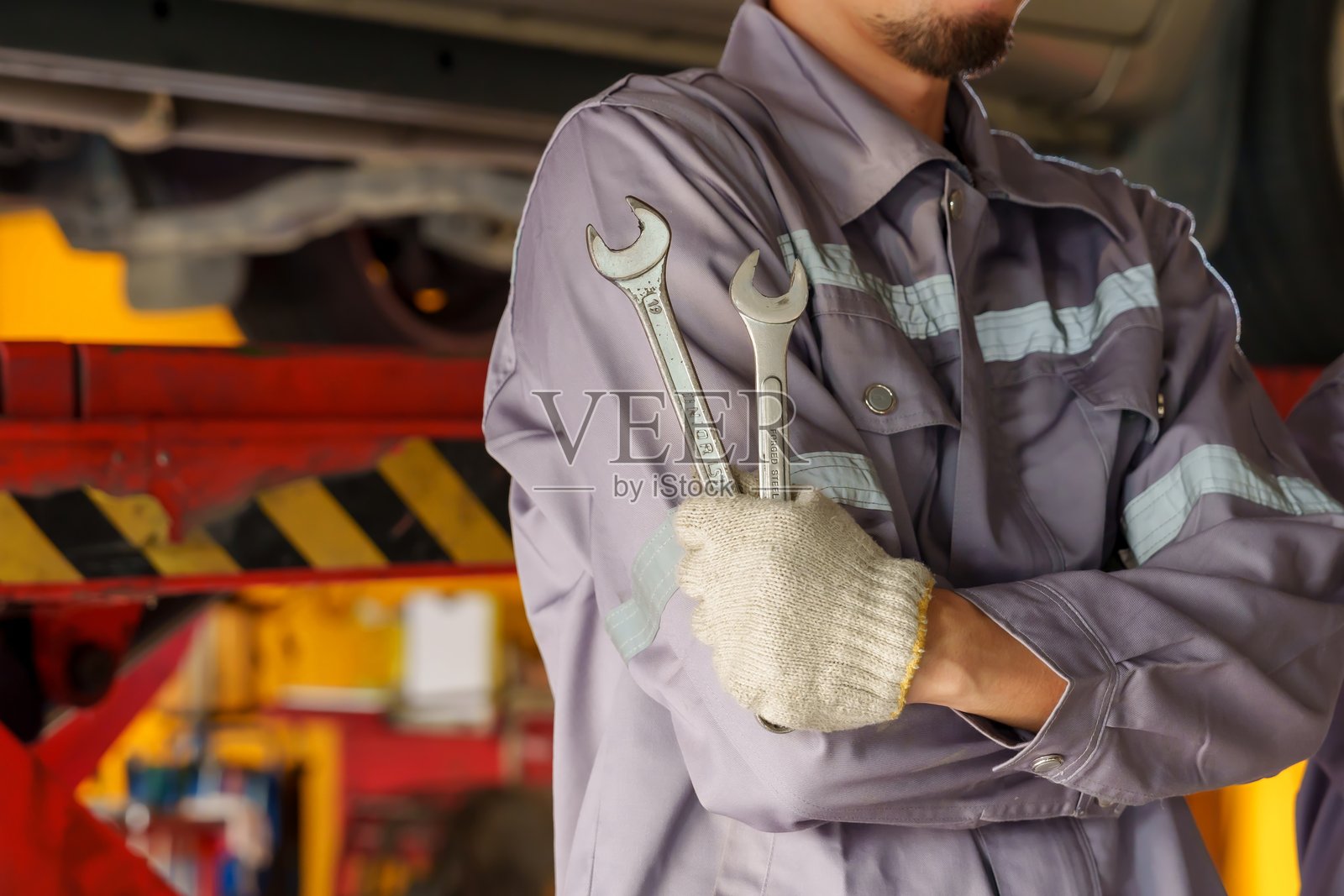 机械师特写，双臂交叉，胸前夹着两把扳手。穿着带反光条的灰色工作服，展现出力量、自信和汽车维修的专业技能。照片摄影图片