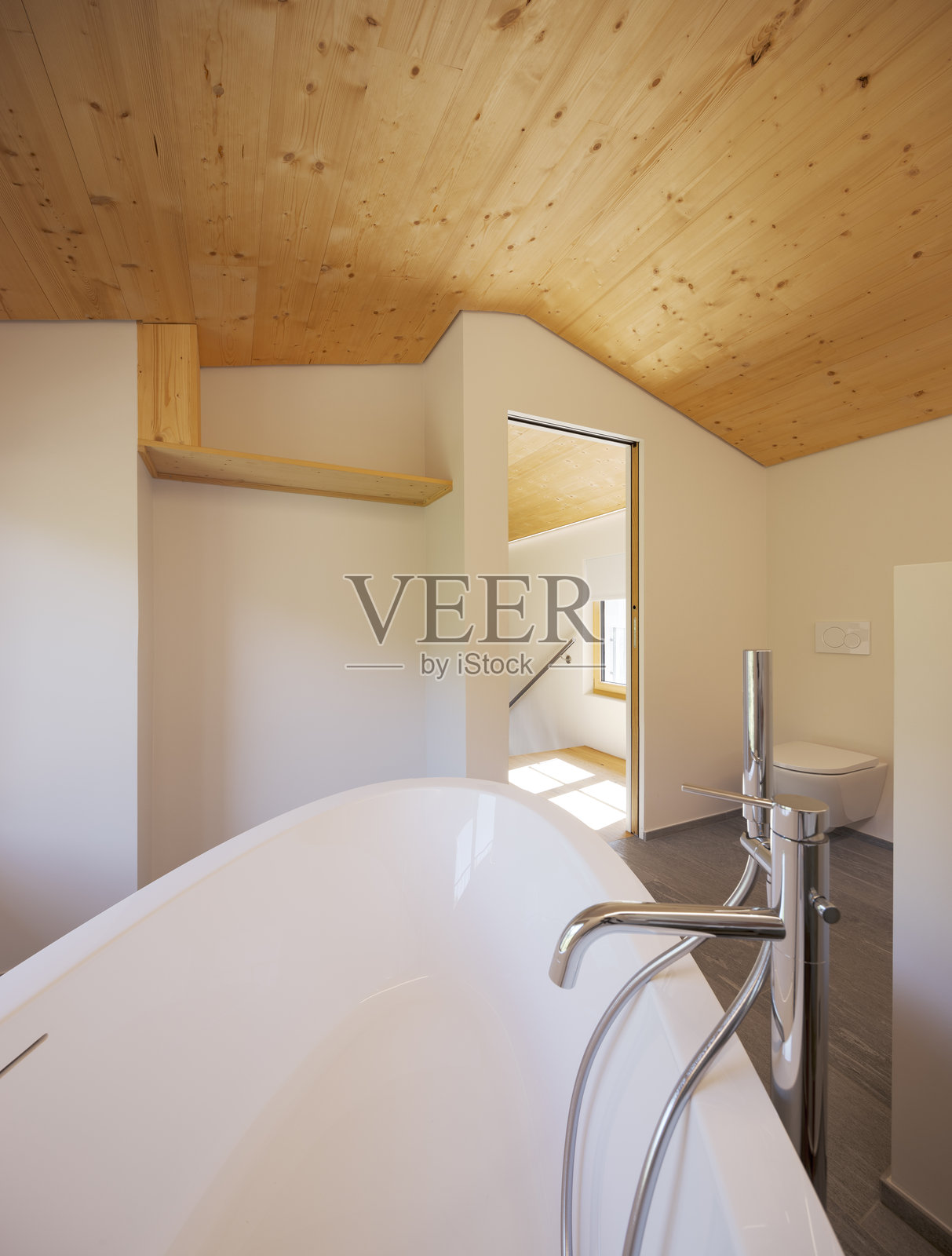 带有不锈钢水龙头的浴缸细节，屋顶是倾斜的木质结构。门是敞开的，可以隐约看到窗户和楼梯栏杆。照片摄影图片
