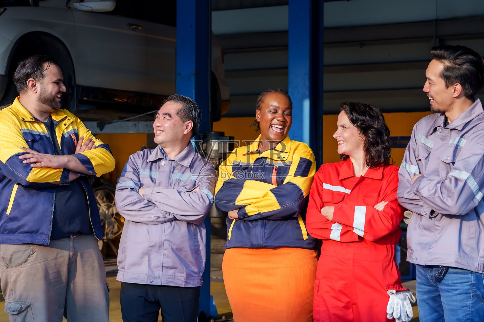 一群多元化的机械师站在一起，双臂交叉，微笑着，身穿色彩鲜艳的工作服，展现出团队合作、友谊和在专业汽车环境中的自信。照片摄影图片