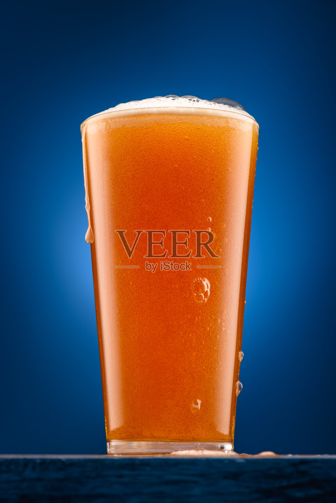 一杯啤酒放在木制支架上，背景为深蓝色，啤酒正倒入杯中，物体的特写，啤酒泡沫丰富，气泡翻腾。照片摄影图片