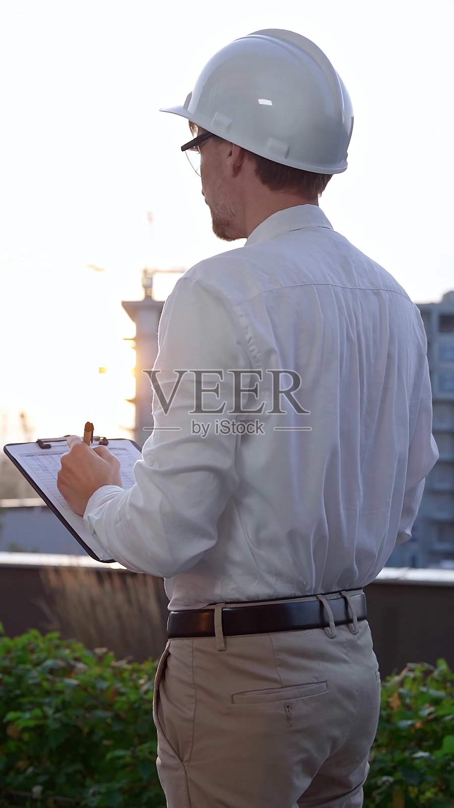 一名穿着白衬衫和安全帽的土木工程师在日出时分检查建筑工地，背面和垂直视角记录在夹板上的笔记。建筑师在工作。照片摄影图片