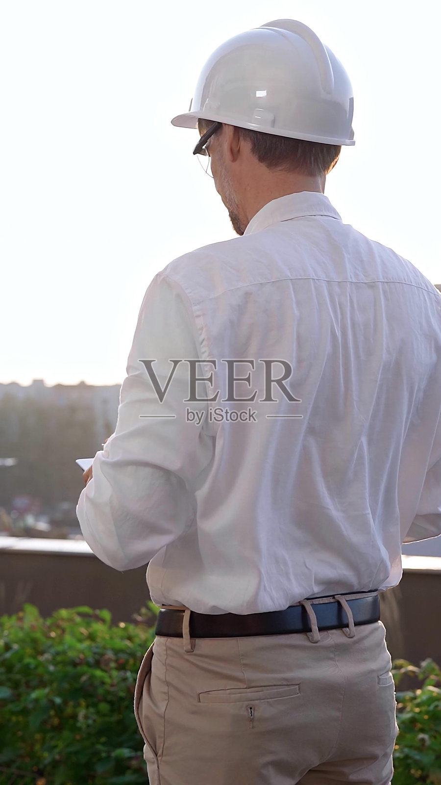 一名男性建筑工程师穿着白衬衫和安全帽，在日出时分检查建筑工地，使用数字平板电脑，背面和垂直视角。建筑概念照片摄影图片