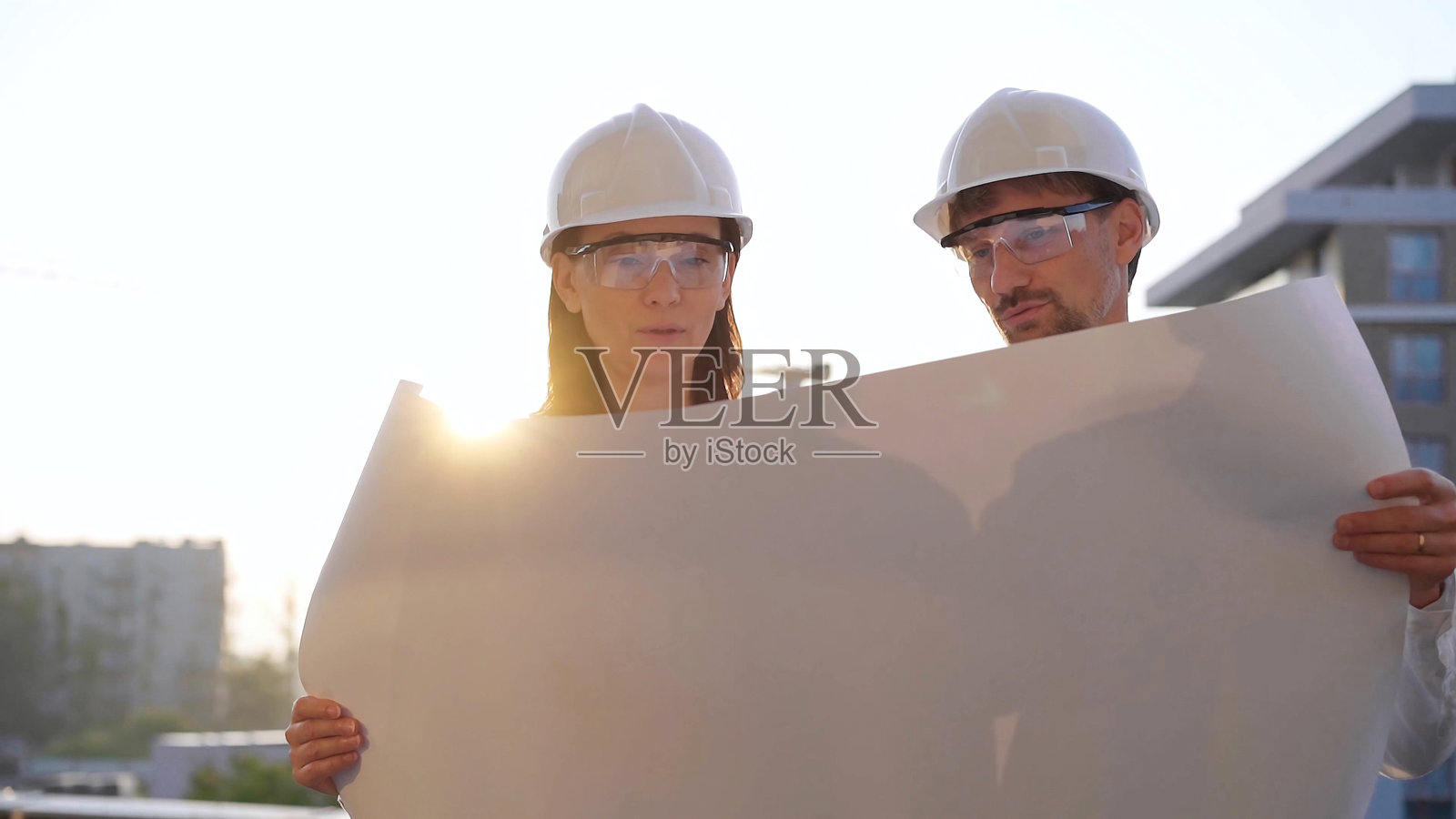 男女建筑师佩戴安全头盔和眼镜，在日落时分的建筑工地前方查看蓝图。照片摄影图片