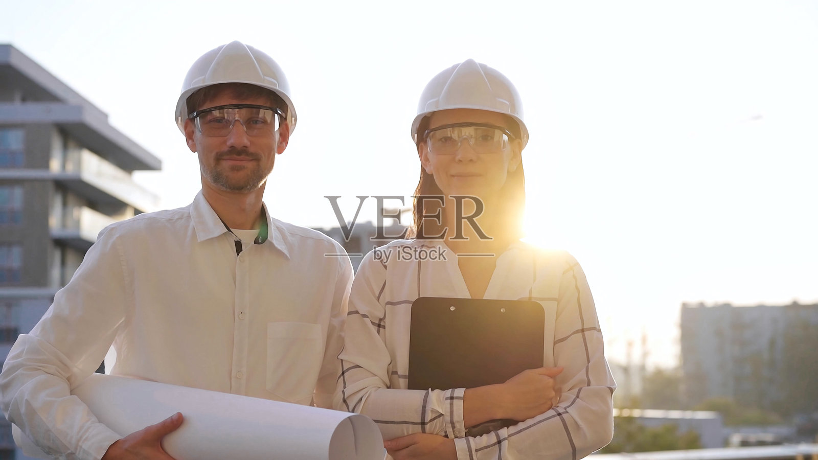 男女建筑师佩戴安全头盔，站在一起，面向镜头，背景是日出或日落时的建筑工地。建筑与工程概念。照片摄影图片
