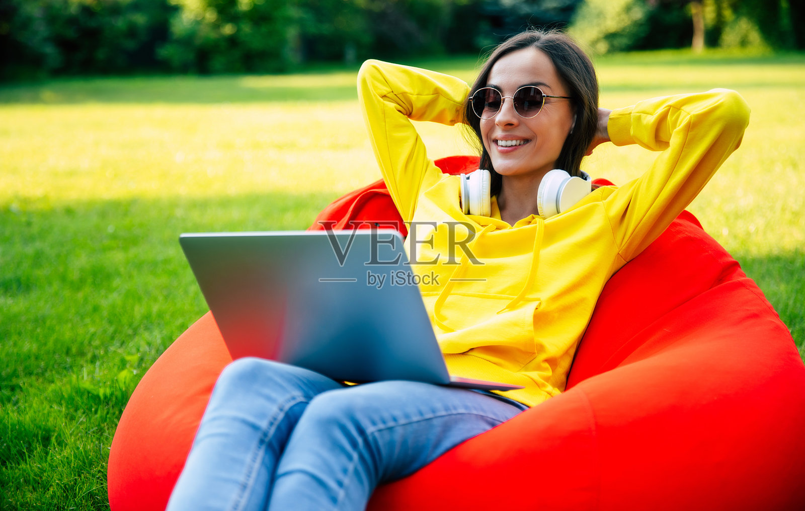 现代微笑自信的年轻女子的全身形象，穿着休闲服，戴着太阳镜，坐在绿色草坪花园的豆袋椅上，拿着笔记本电脑和手机。女性自由职业者，商业，工作照片摄影图片