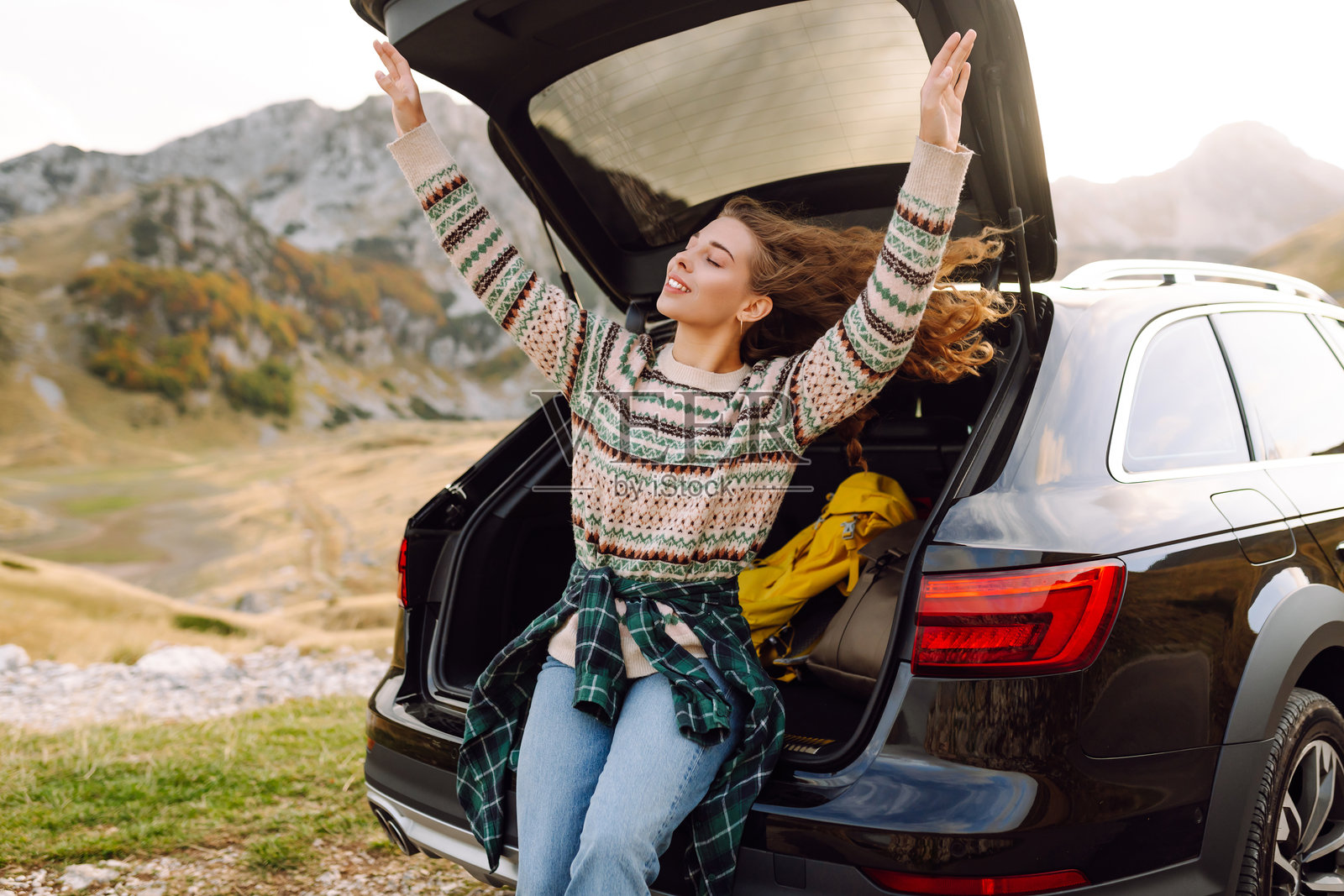 年轻女子坐在她的车后备厢里，车停在蜿蜒的道路旁，穿过崎岖的地形。公路旅行。秋季风景。照片摄影图片