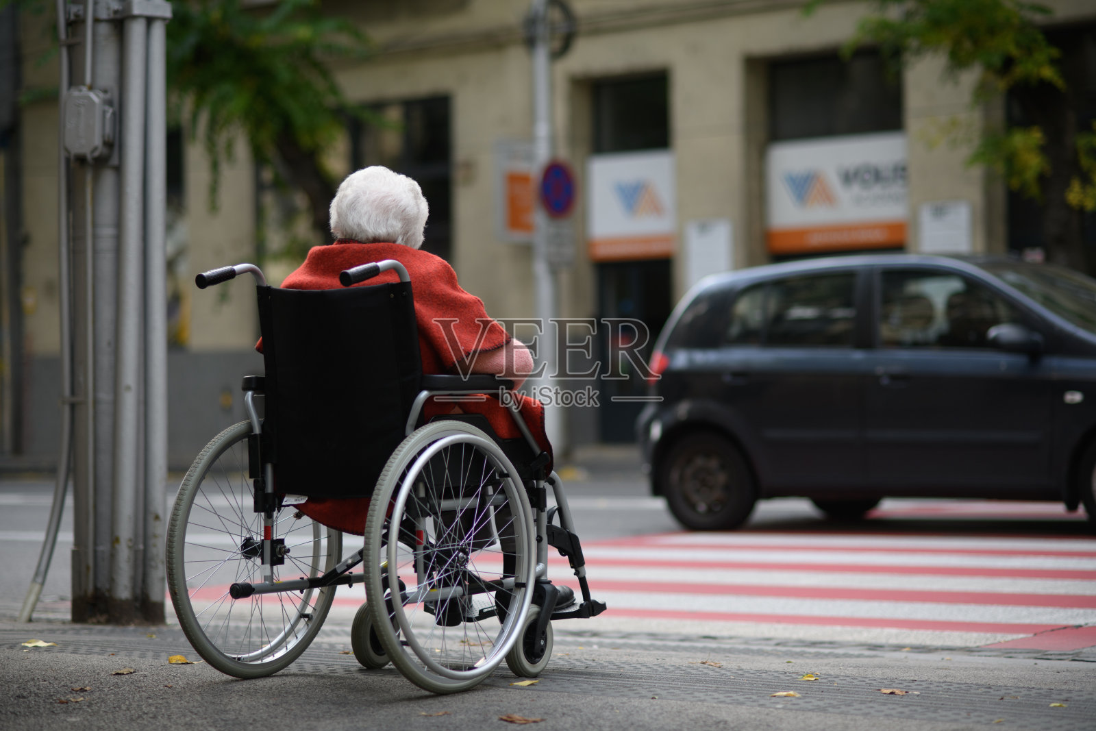 坐在轮椅上的残疾老年女性在十字路口等待。背影。照片摄影图片