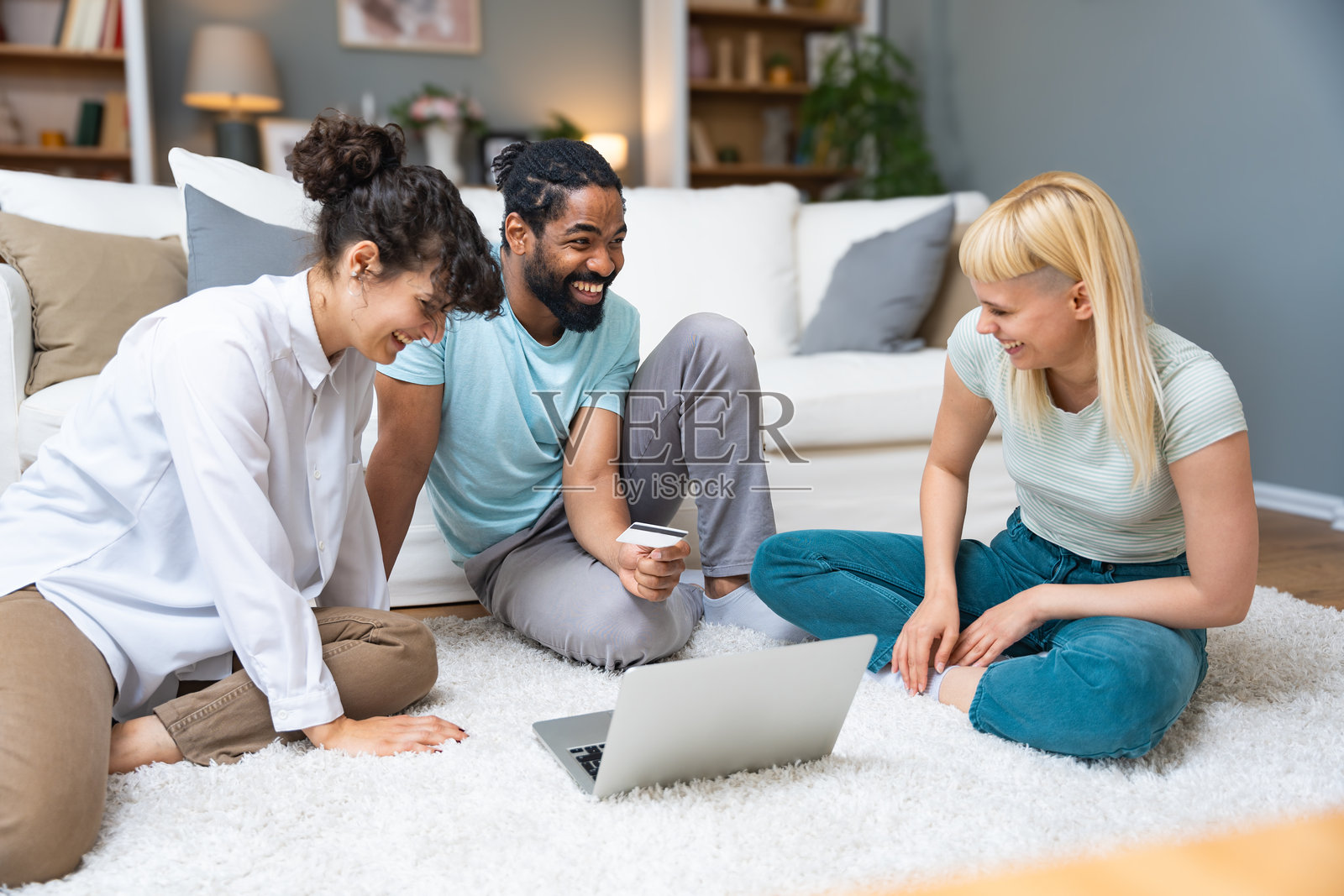 三位兴奋的朋友在家里的客厅地板上用笔记本电脑和信用卡在线购买度假票。人们在网上购物和下单。照片摄影图片