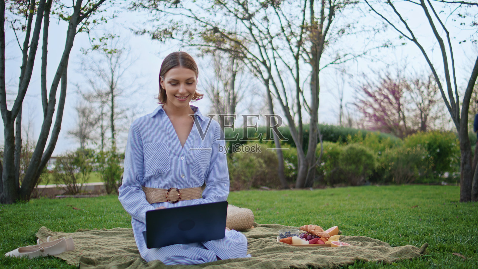坐在野餐毯上的自由职业者，使用笔记本电脑吃零食，微笑的棕发女性。照片摄影图片