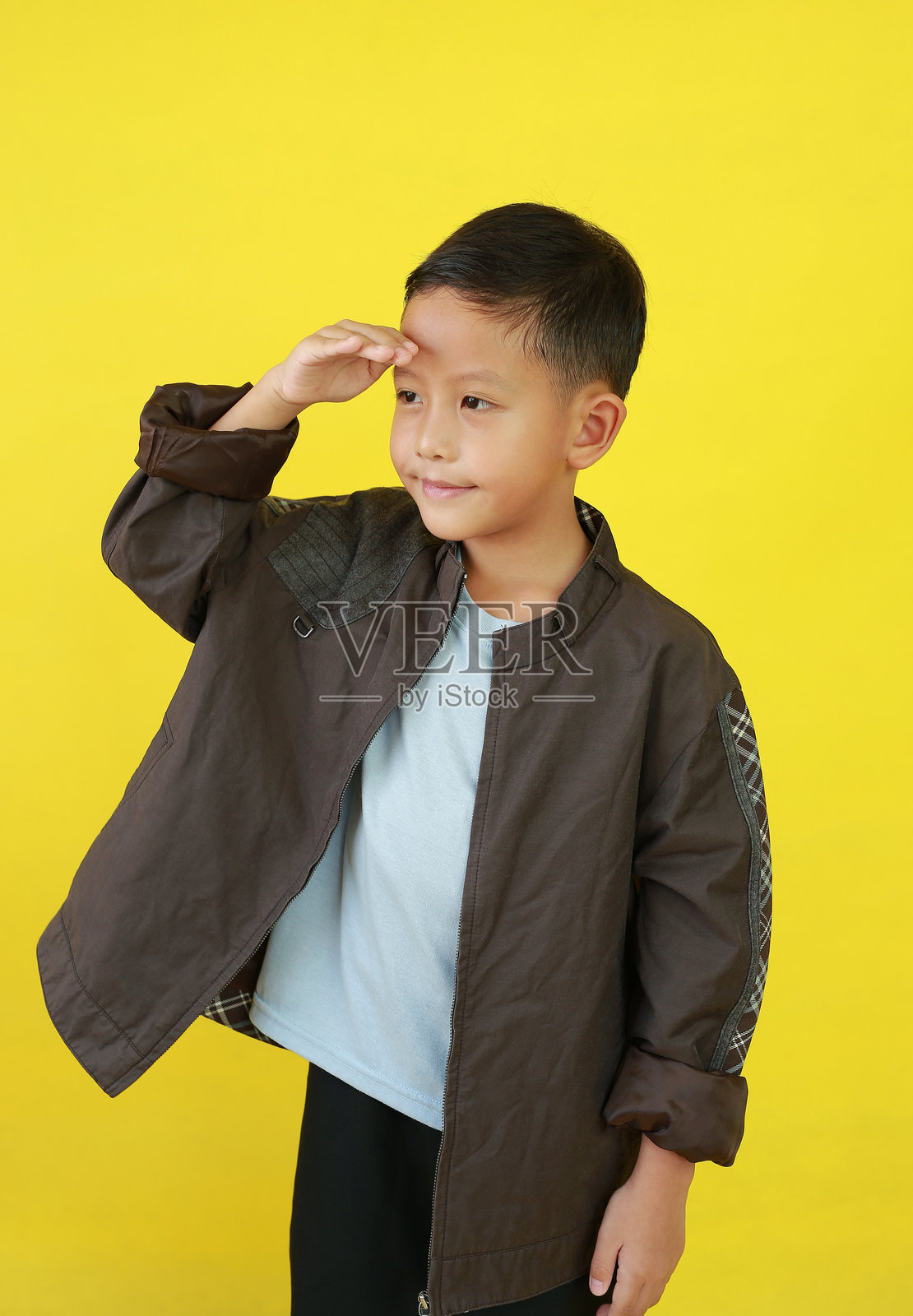 微笑的亚洲男孩穿着夹克，手放在额头上，目光投向一旁，背景为黄色的摄影棚。照片摄影图片