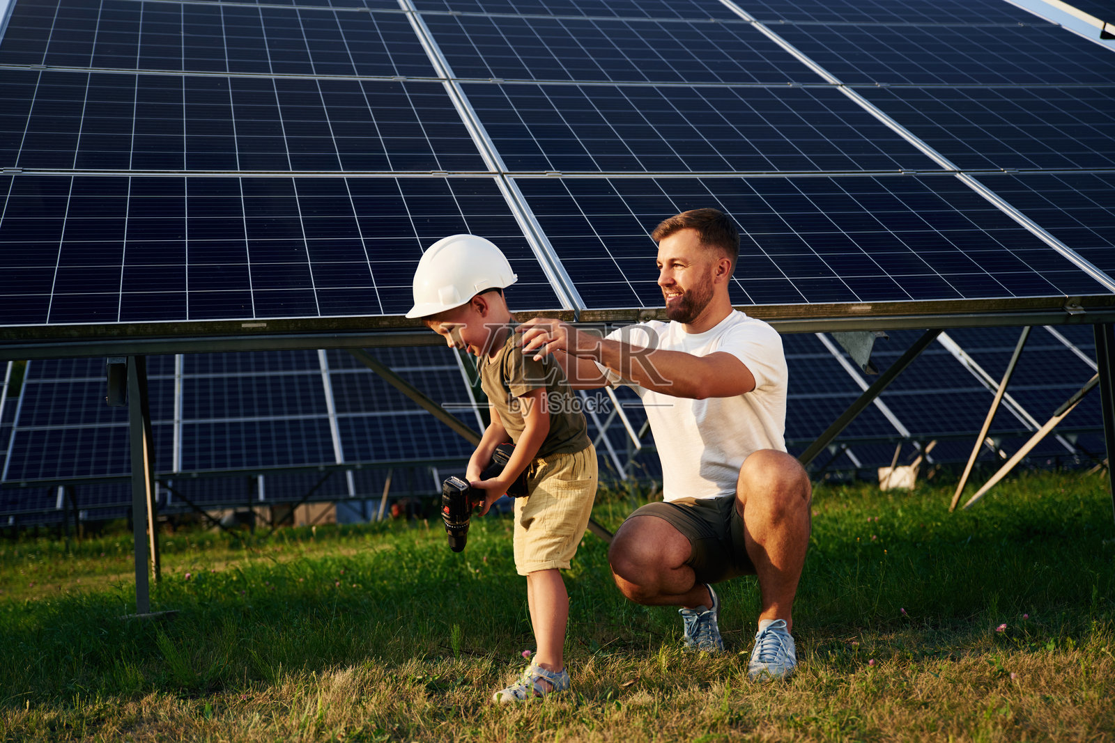 戴着安全帽，手持电钻的父亲与他的小儿子在太阳能电池板旁一起。照片摄影图片