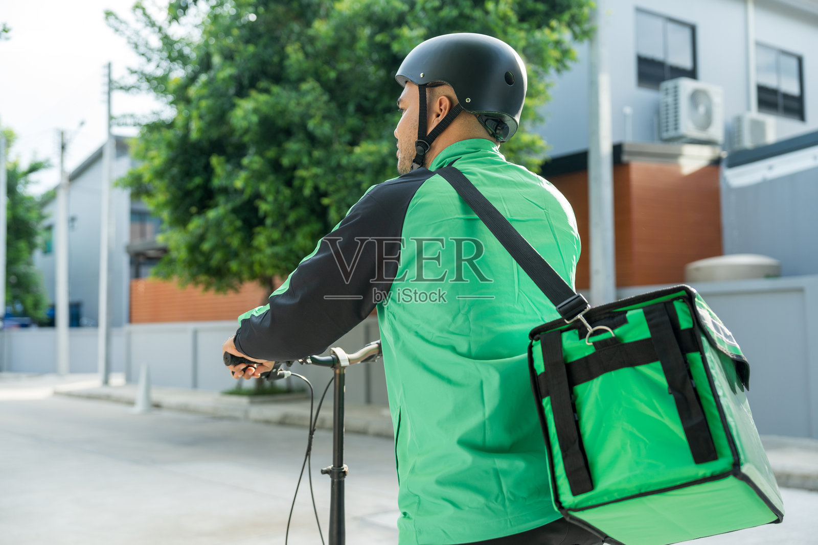 骑着自行车的外卖员，穿着绿色制服和头盔，正在为顾客送餐。快乐的外卖员背着绿色背包，将货物送给顾客。照片摄影图片