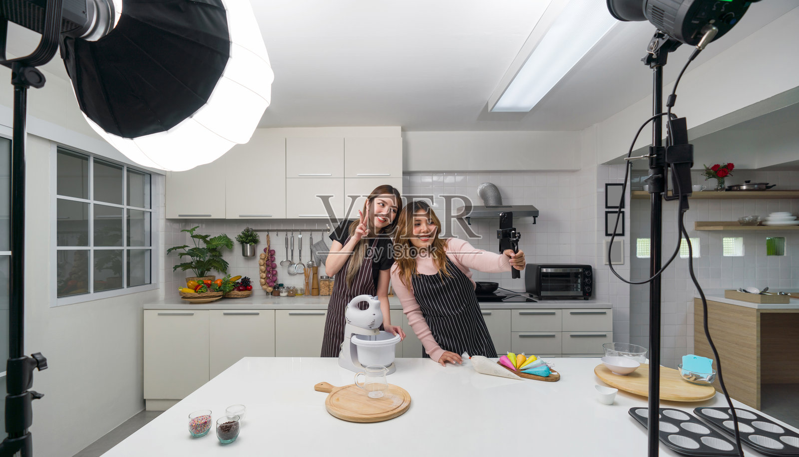两位穿着围裙的亚洲女性在厨房里微笑着做饭，手持自拍杆上的智能手机和摄影灯光设备，正在为社交博主录制视频博客。照片摄影图片