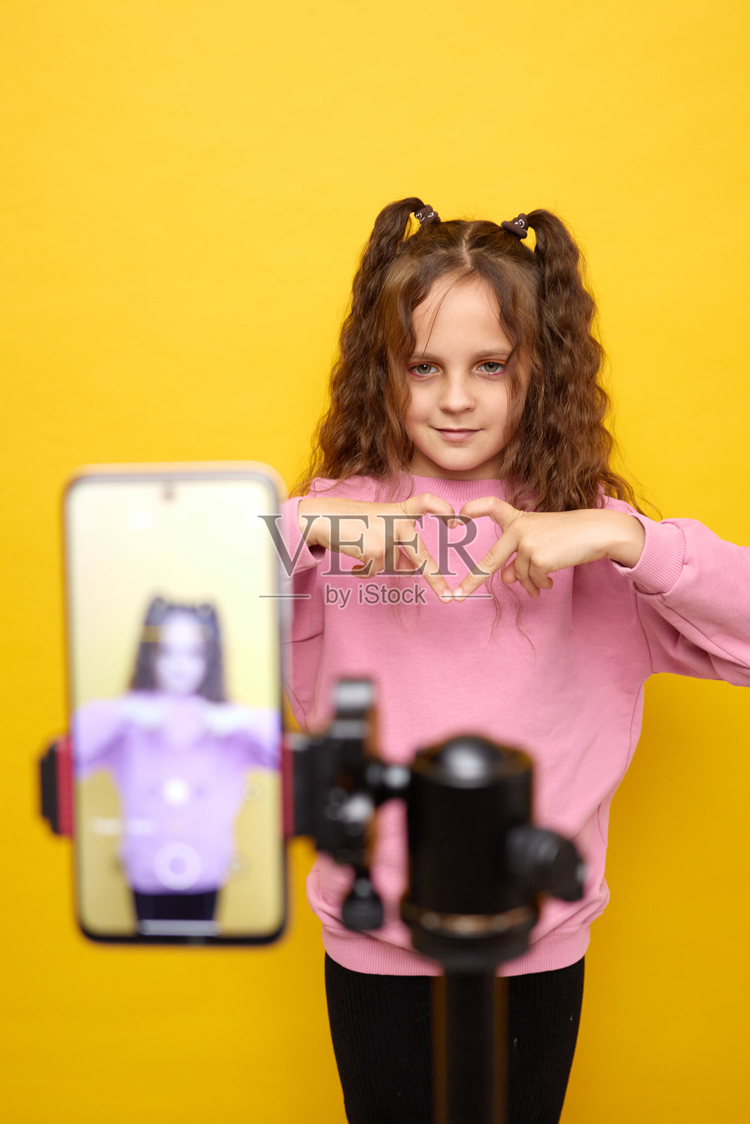可爱的女孩在录制时尚潮流的 vlog，化着妆在工作室里跳舞，用手机表达自己，双手比心，背景为黄色。照片摄影图片