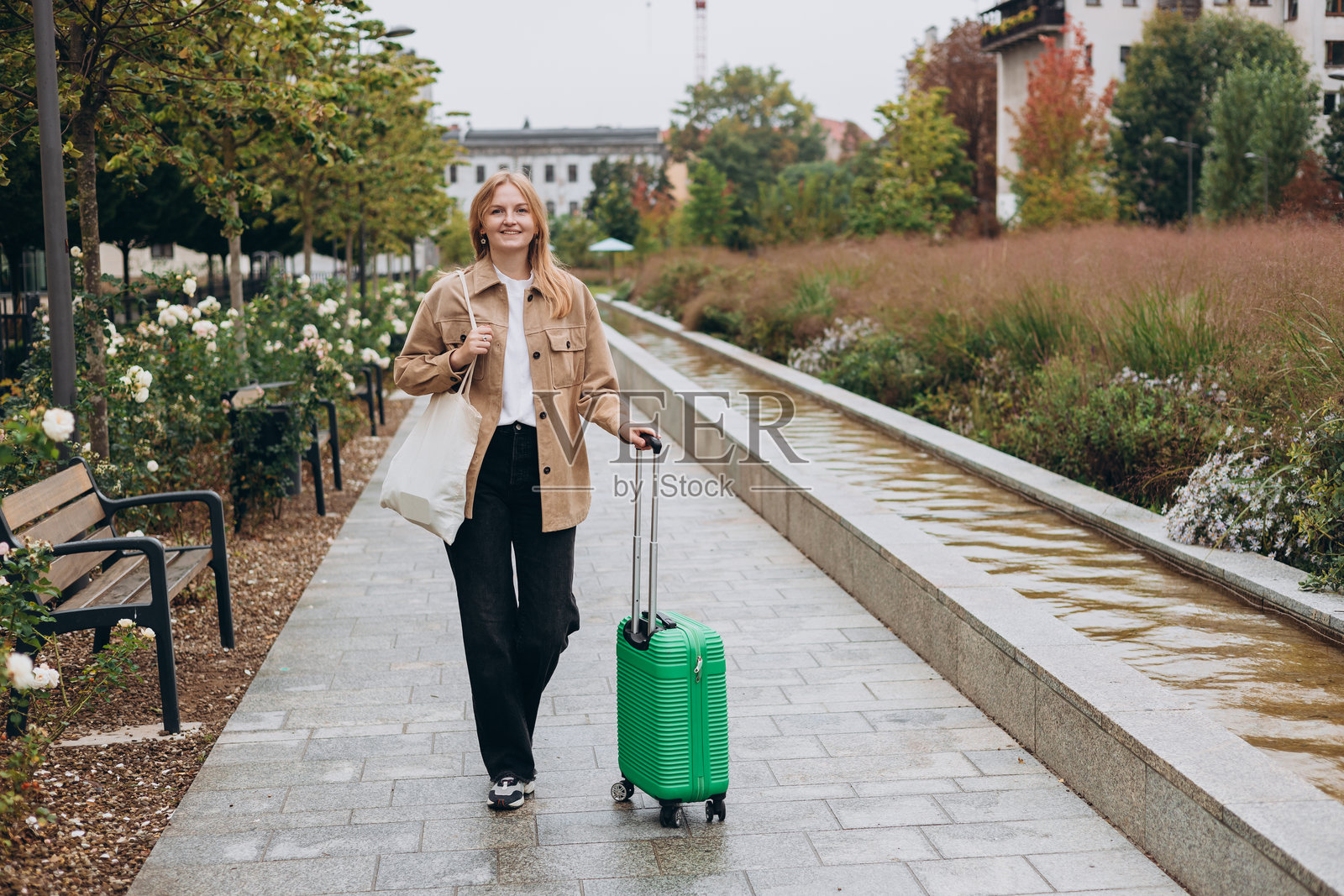 穿着休闲服的年轻女性在城市公园里愉快地走着，拖着绿色行李箱。30多岁的女性在城市背景下拖着行李箱。旅行时尚。全身照。照片摄影图片