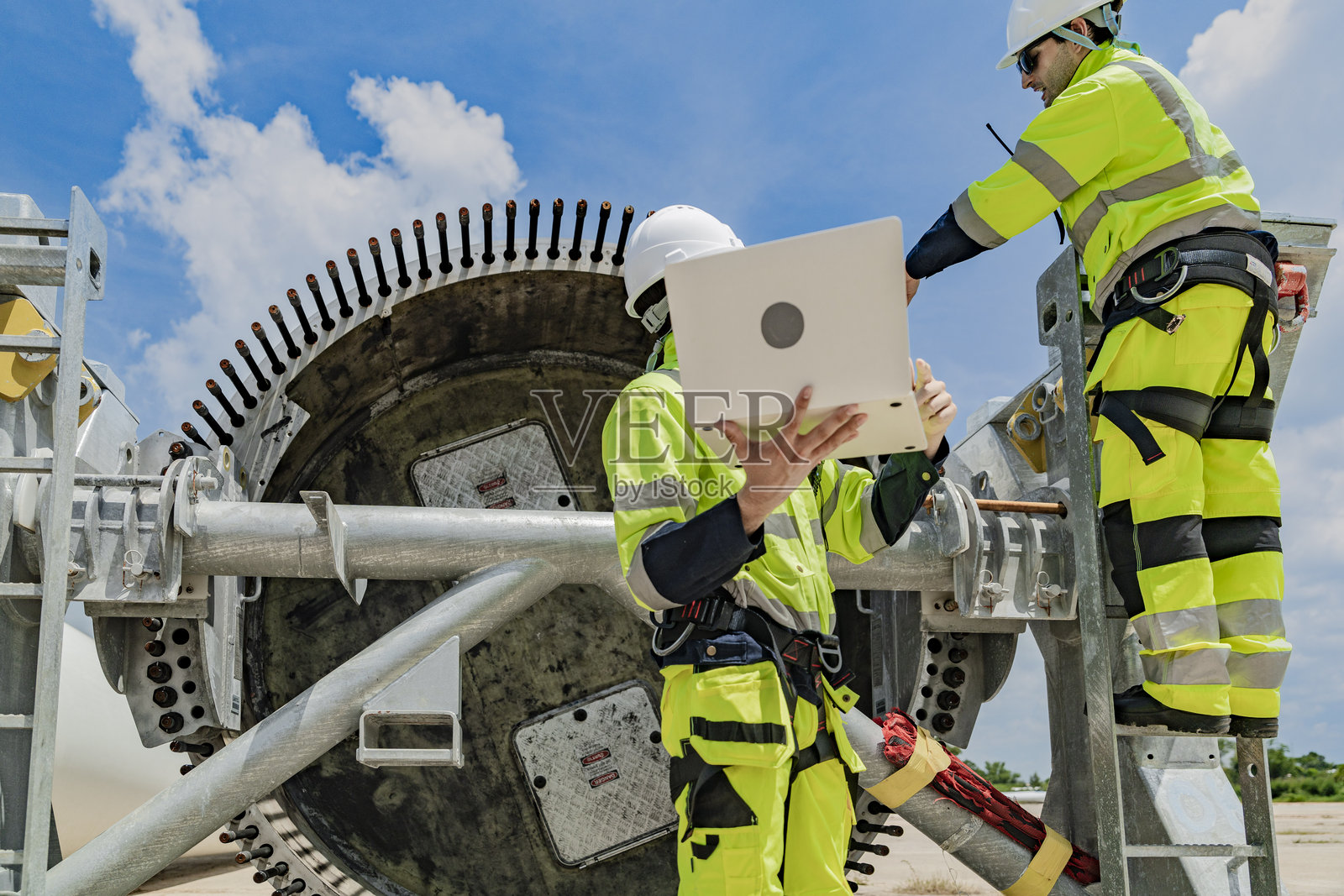 两名工程师在风力涡轮机组件上工作，一人使用笔记本电脑，另一人在梯子上检查零件。可再生能源技术、风能项目以及能源行业的团队合作。照片摄影图片