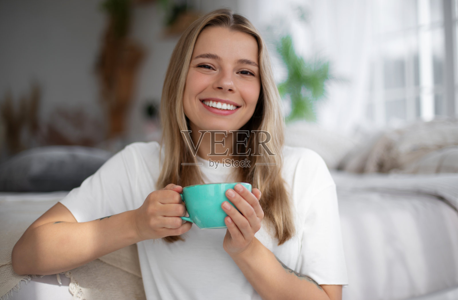 一位年轻女性盘腿坐在地板上，微笑着手握一杯。她身处一个明亮舒适的卧室，柔软的床上用品、自然光和植物营造出轻松宁静的氛围。照片摄影图片