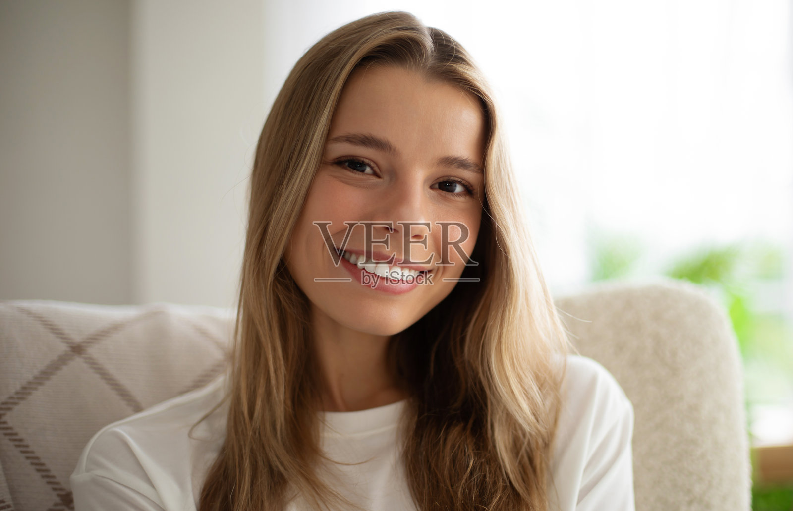 一位迷人的年轻女性微笑着坐在室内，长长的金发披肩，穿着休闲的白色衬衫。背景明亮而舒适，配有白色椅子和柔和的自然光线。照片摄影图片