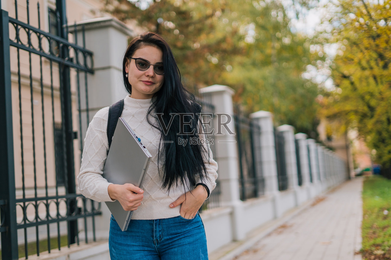 一位微笑的女学生站在教职员工面前，手里拿着笔记本电脑，背上背着书包。秋天的日子，色彩斑斓，她散发着快乐和热情的气息。照片摄影图片