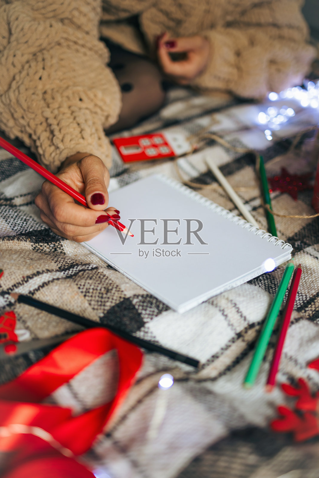 穿着针织毛衣的女人坐在温暖的格子毯子上，旁边有圣诞玩具和礼品盒，正在笔记本上写下她的新年目标。照片摄影图片