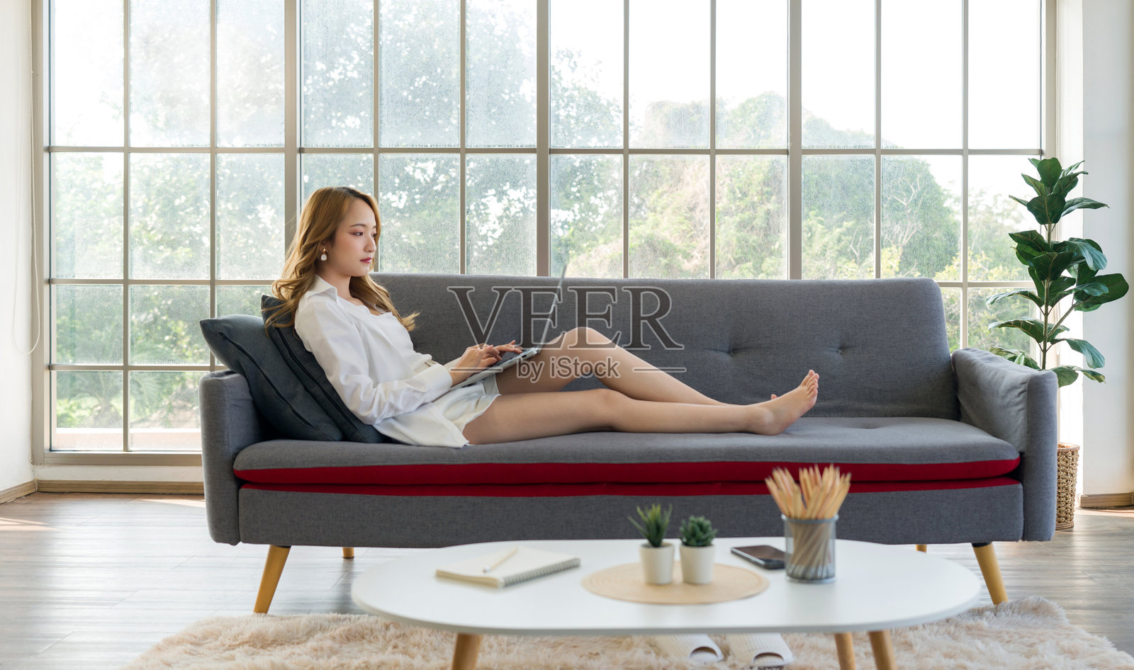 年轻的亚洲女性穿着白衬衫和短裤，坐在沙发上，轻松地在笔记本电脑键盘上打字，房间明亮，窗户很大。照片摄影图片