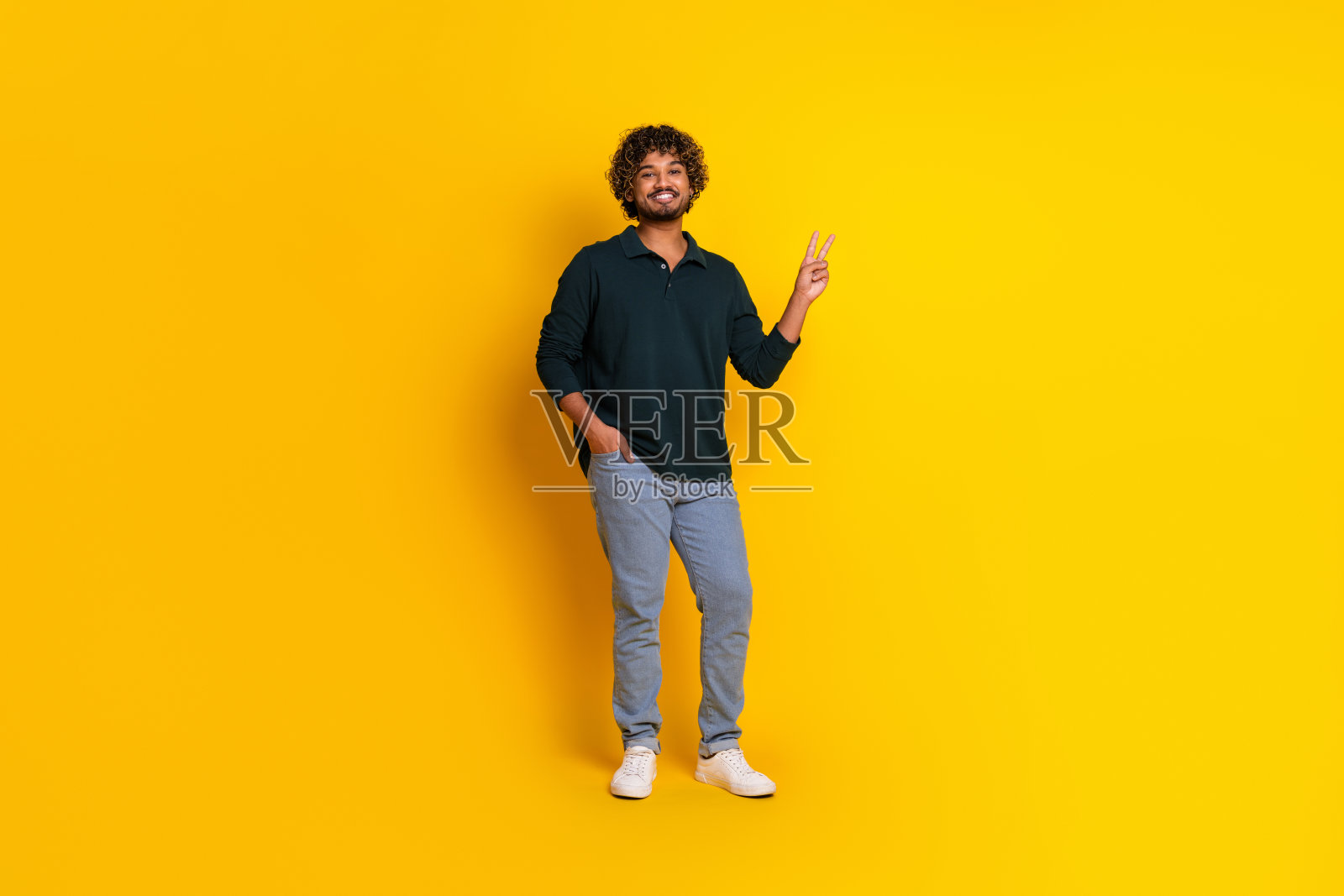 年轻男子在鲜艳的黄色背景前摆出和平手势，穿着休闲的黑色衬衫和牛仔裤。照片摄影图片