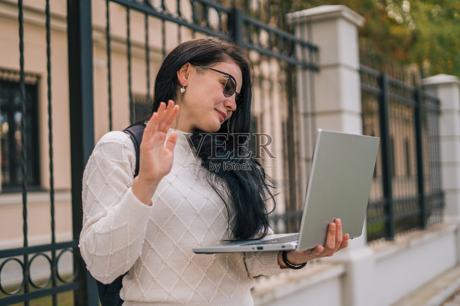 一位微笑着、漂亮的女性穿着舒适的毛衣，在街上手持笔记本电脑，正在进行视频通话并向镜头挥手。照片摄影图片