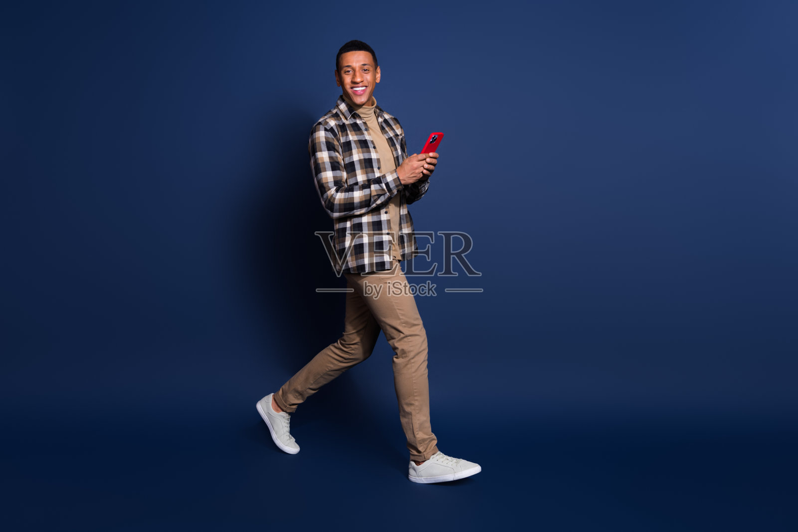 穿着格子衬衫、心情愉快的帅气男士在蓝色背景下使用苹果iPhone和三星现代电子产品打字。照片摄影图片