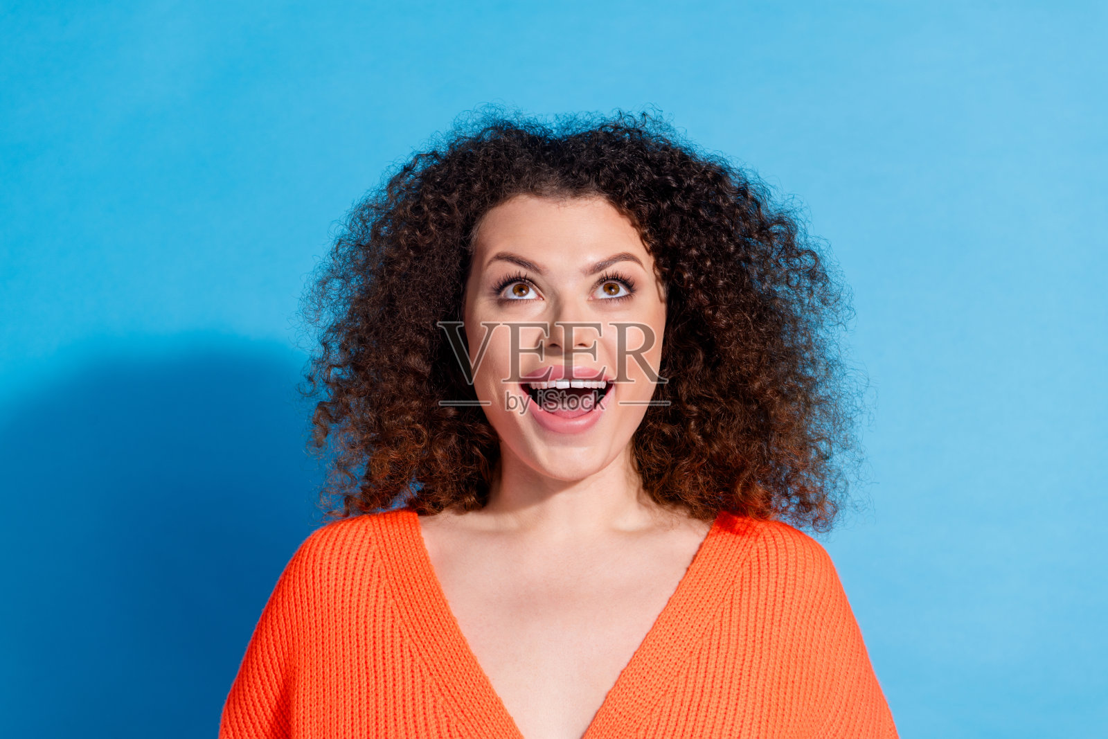 穿着橙色开衫的波浪卷发惊讶女性肖像，注视着蓝色背景上方。照片摄影图片
