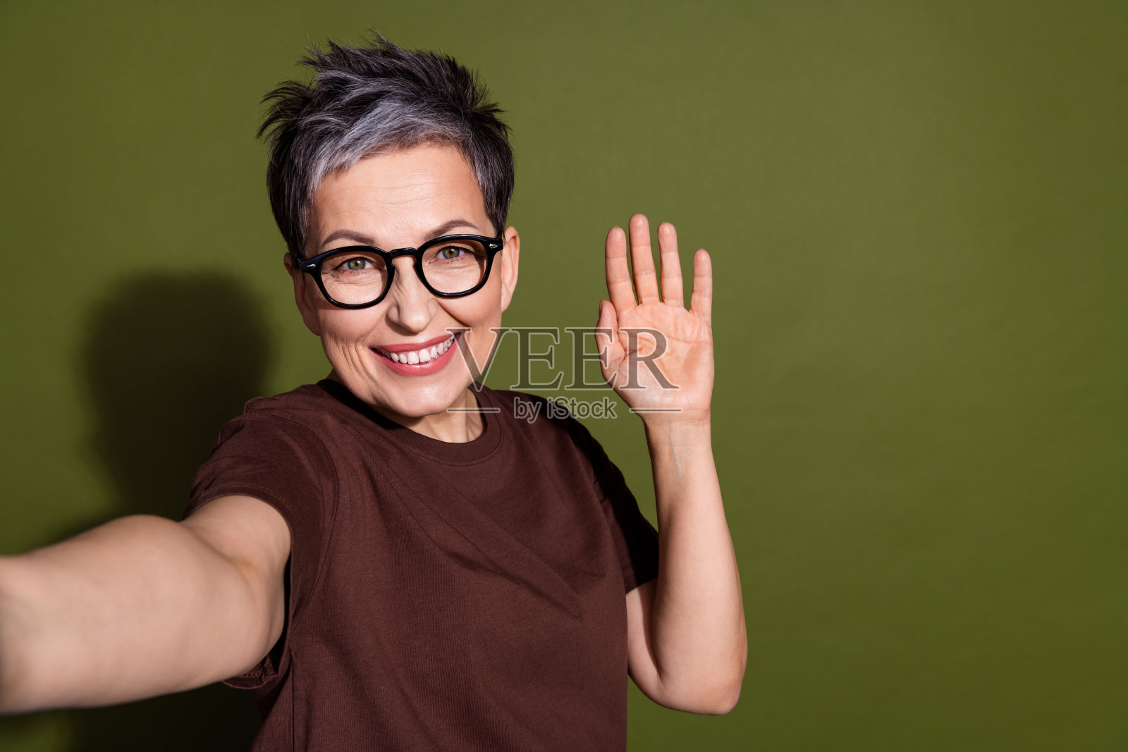 时尚的退休女性在卡其色背景下穿着棕色衣服，微笑着自拍并挥手。照片摄影图片