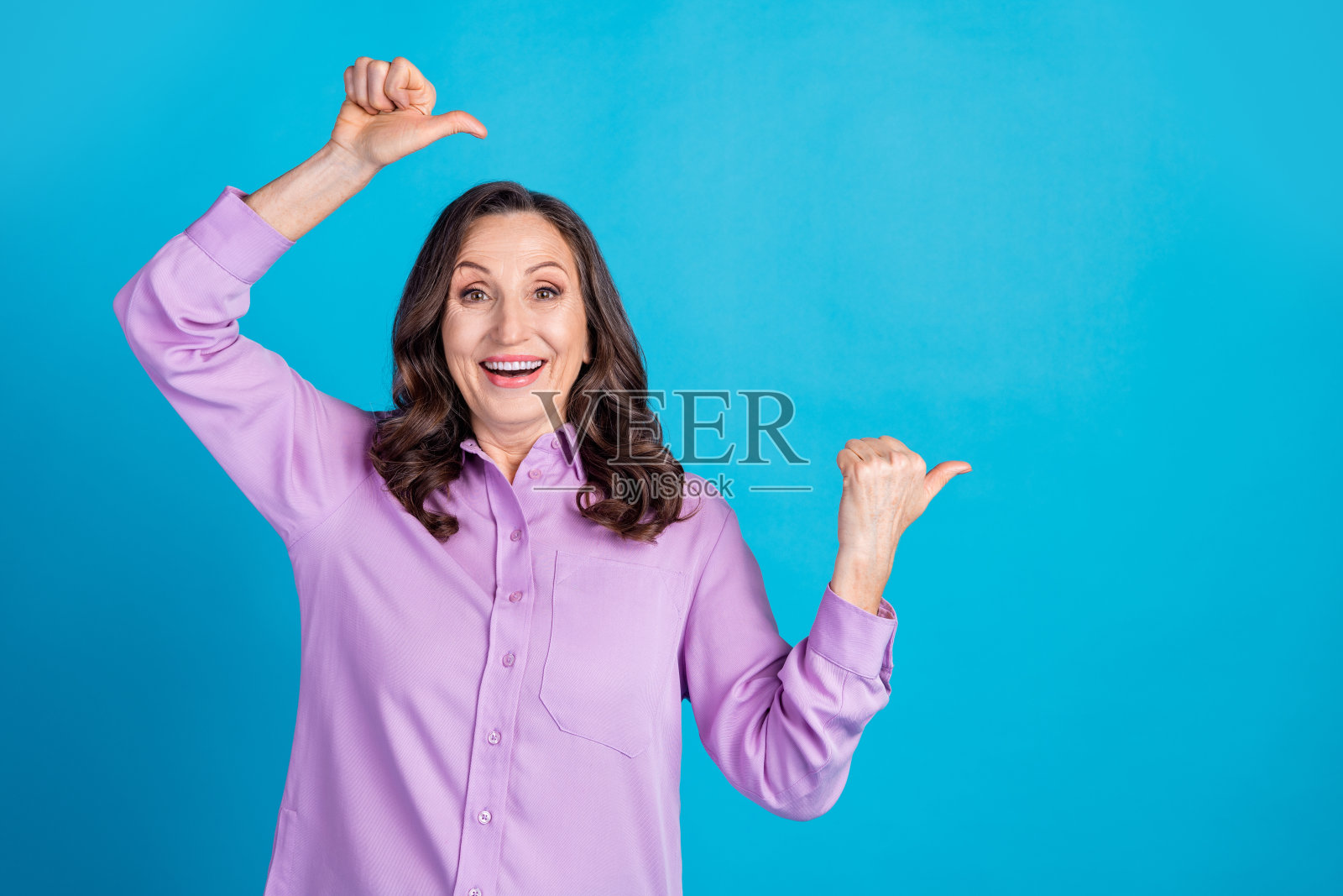 可爱的女孩穿着紫色衬衫，双手竖起大拇指指向空白空间，背景为蓝色。照片摄影图片
