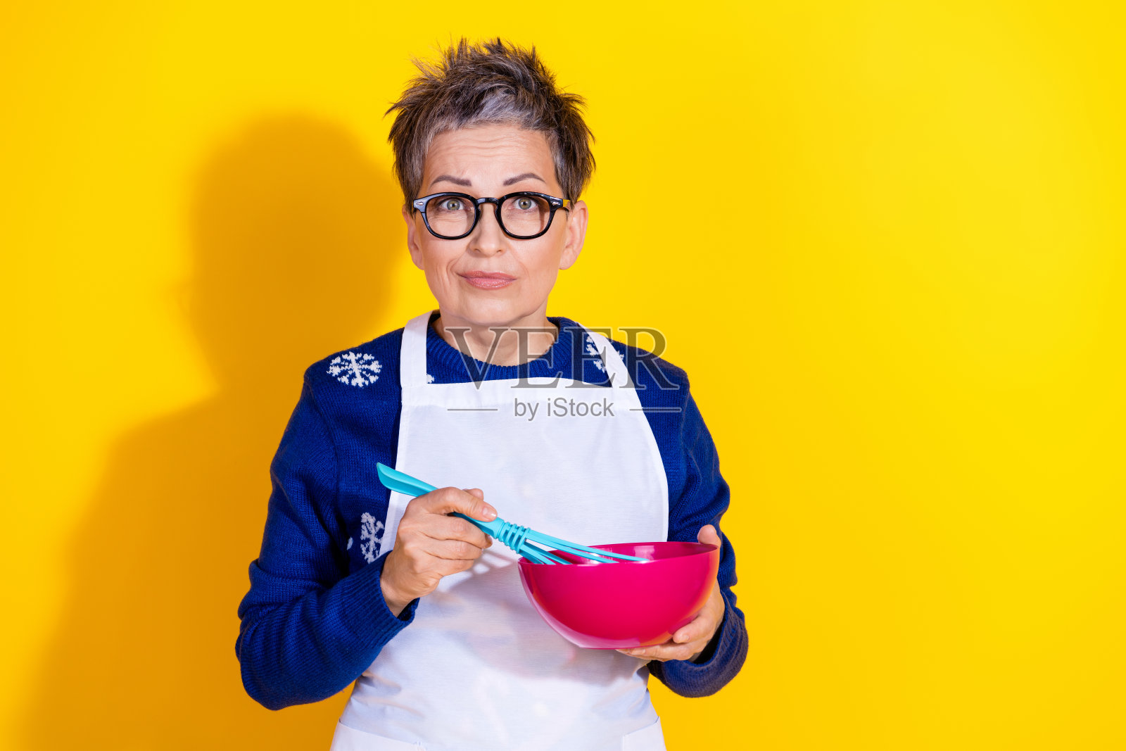 可爱的退休女性穿着毛衣，手持塑料搅拌碗正在混合面团，背景为黄色。照片摄影图片