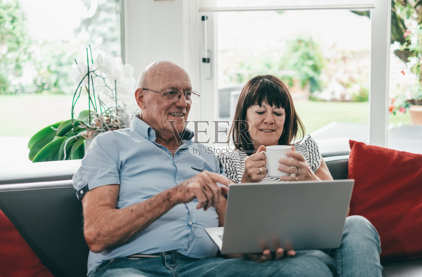 微笑的老年夫妇舒适地坐在沙发上，手里拿着笔记本电脑和茶杯，两个宁静的退休老人享受着家中的宁静时光。照片摄影图片
