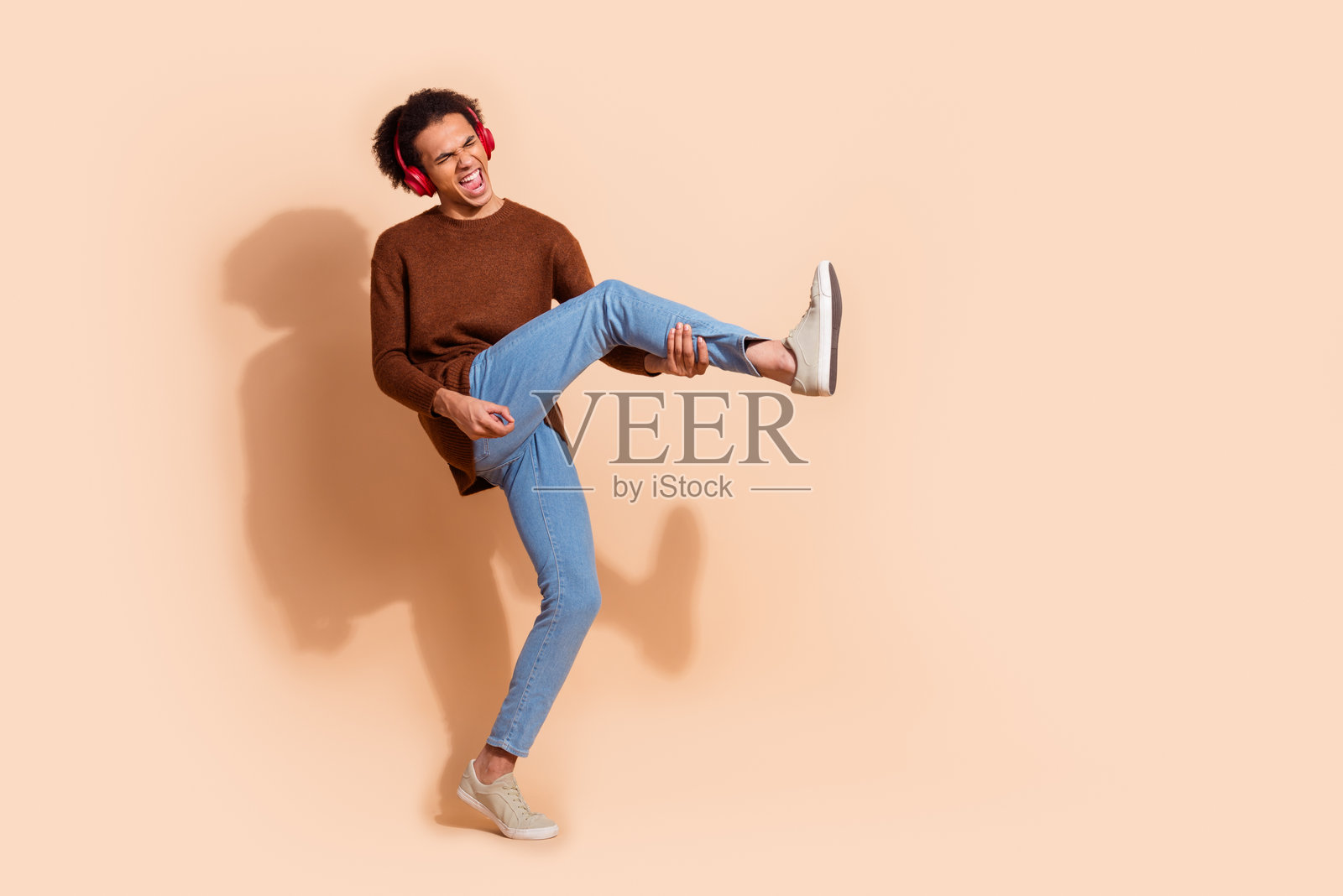 年轻男子穿着休闲服装，在米色背景前顽皮地摆姿势，身穿棕色毛衣和牛仔裤。照片摄影图片