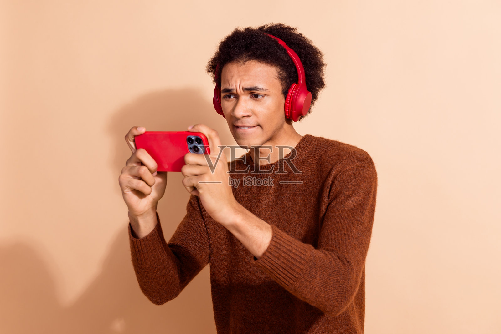 年轻的非裔美国男性穿着休闲服，专注地用红色手机和耳机玩手机游戏，背景为米色。照片摄影图片