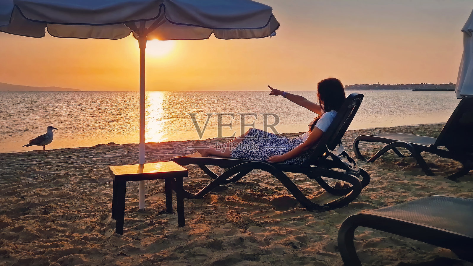 年轻女性坐在海滩的躺椅上，用食指指向地平线上的日出。美丽的女性在海边享受日光浴。夏季假期海滨，度假乐趣的概念。照片摄影图片