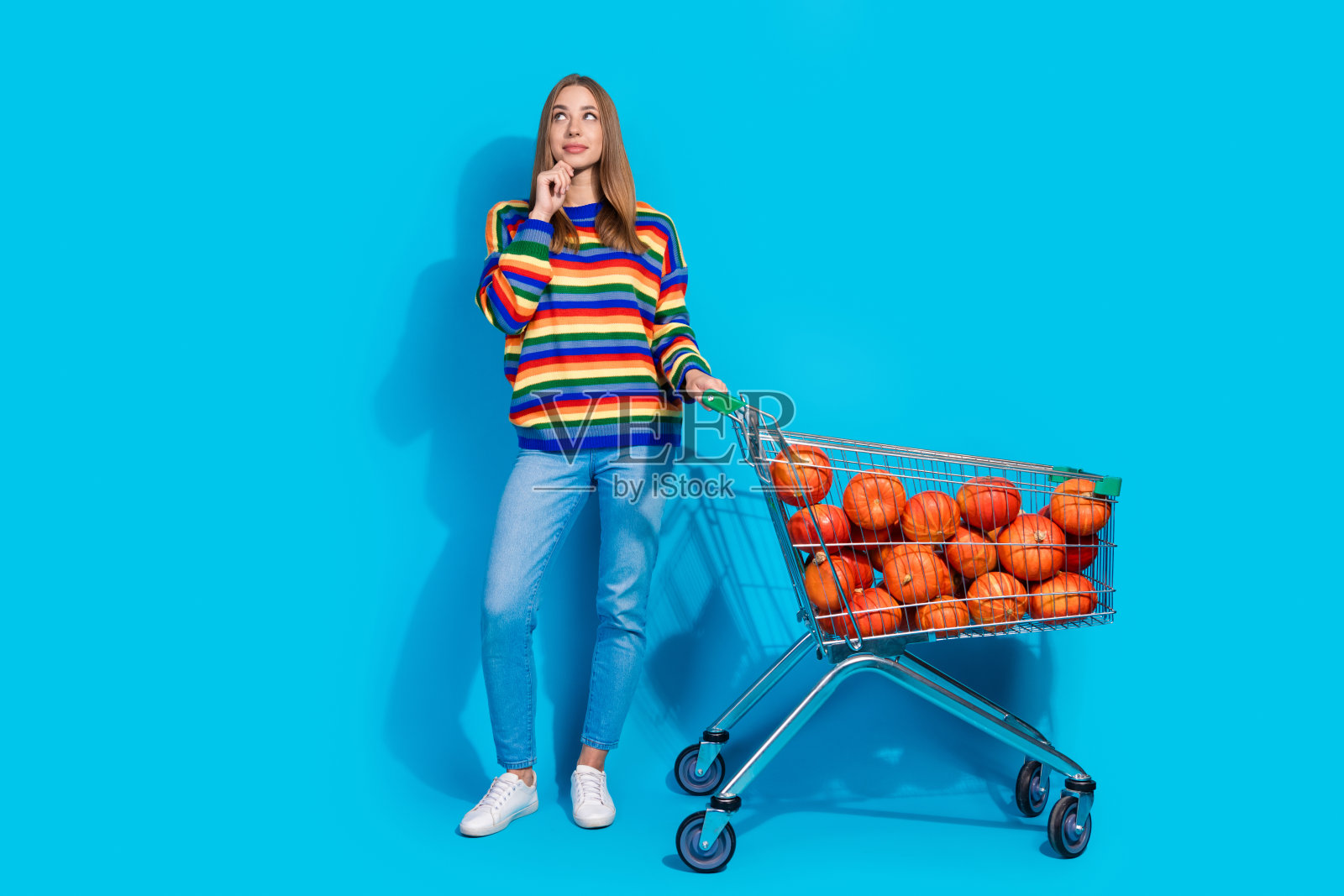 可爱的漂亮女孩穿着条纹彩色衣服，推着装满南瓜的手推车，背景为空旷的蓝色。照片摄影图片